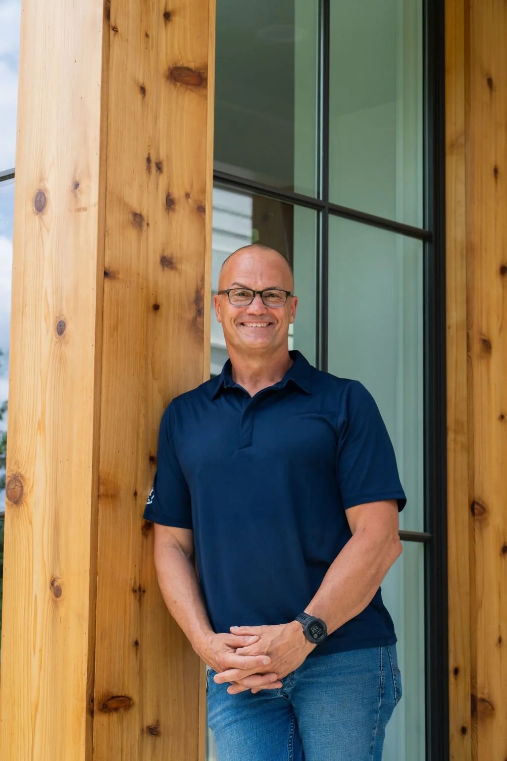 A smiling man with glasses wearing a navy blue polo shirt and jeans standing outdoors next to a wooden structure with large glass windows.