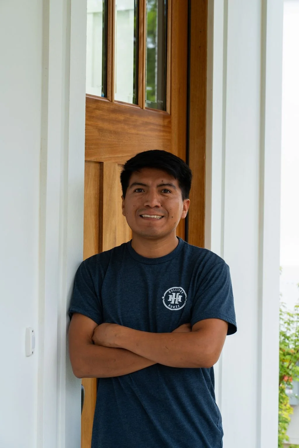 A man with black hair, wearing a dark blue t-shirt, standing with arms crossed in front of a wooden door and glass windows, smiling at the camera.