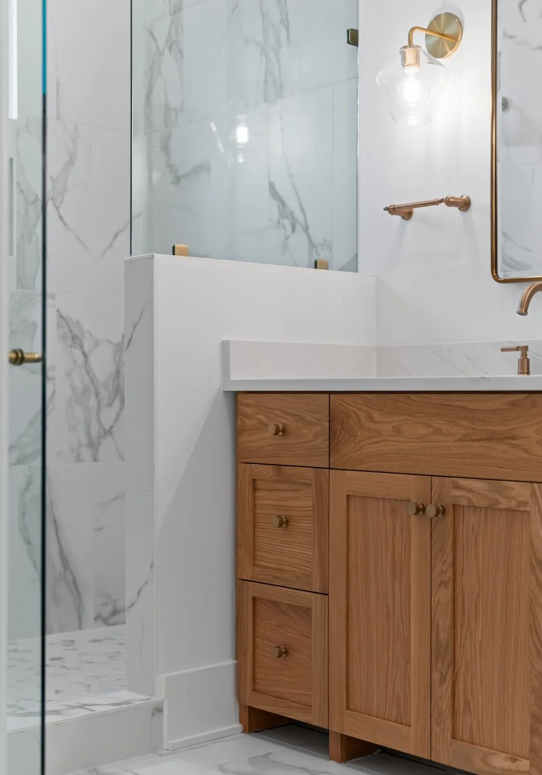 Modern bathroom vanity with wooden cabinets, a white marble countertop, and gold hardware. The wall behind has large marble tiles, with a glass light fixture and a mirror partially visible.