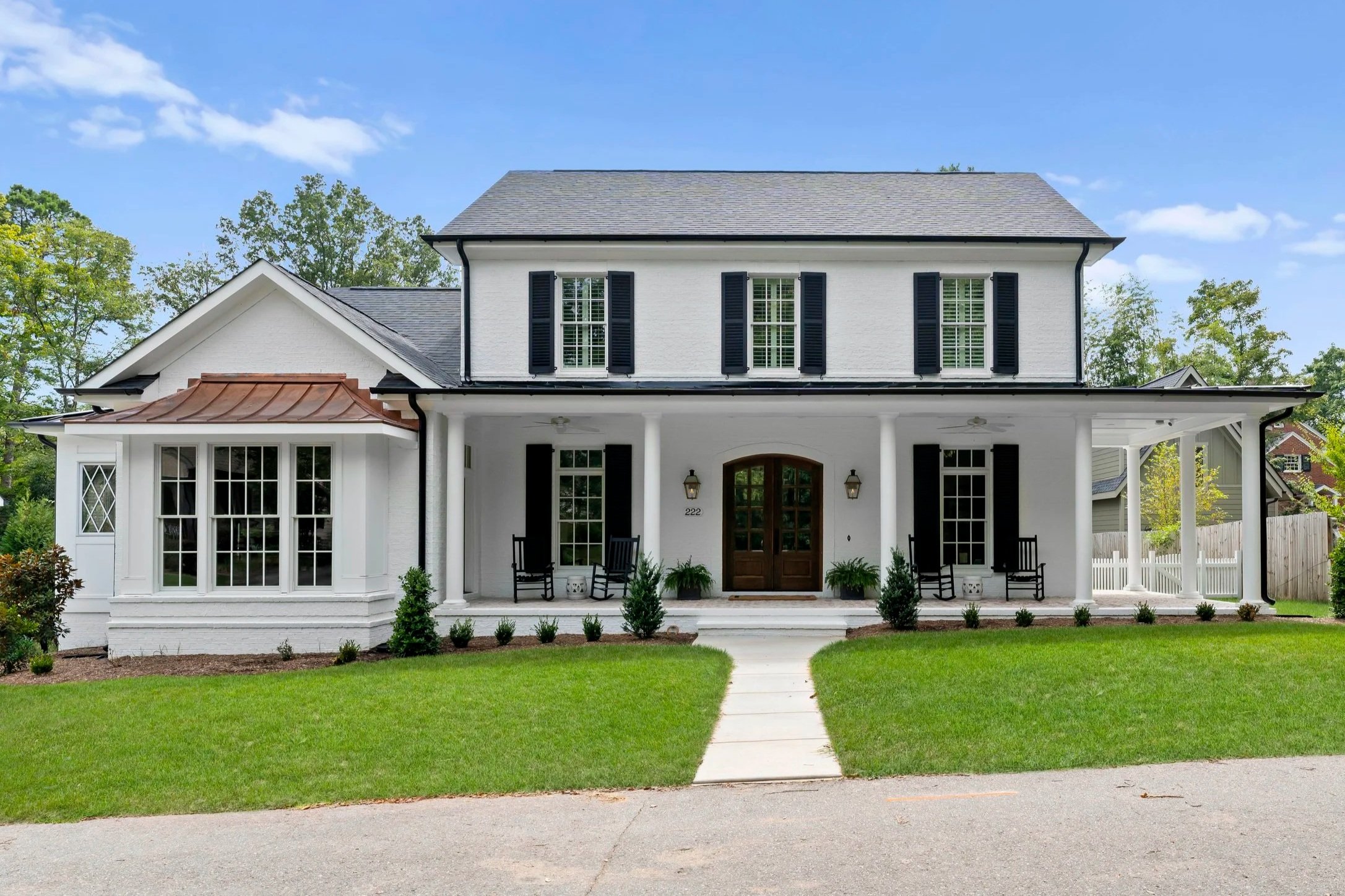 Front view of a two-story white house with black shutters, a covered porch with rocking chairs, and a landscaped lawn with small shrubs and a concrete walkway.