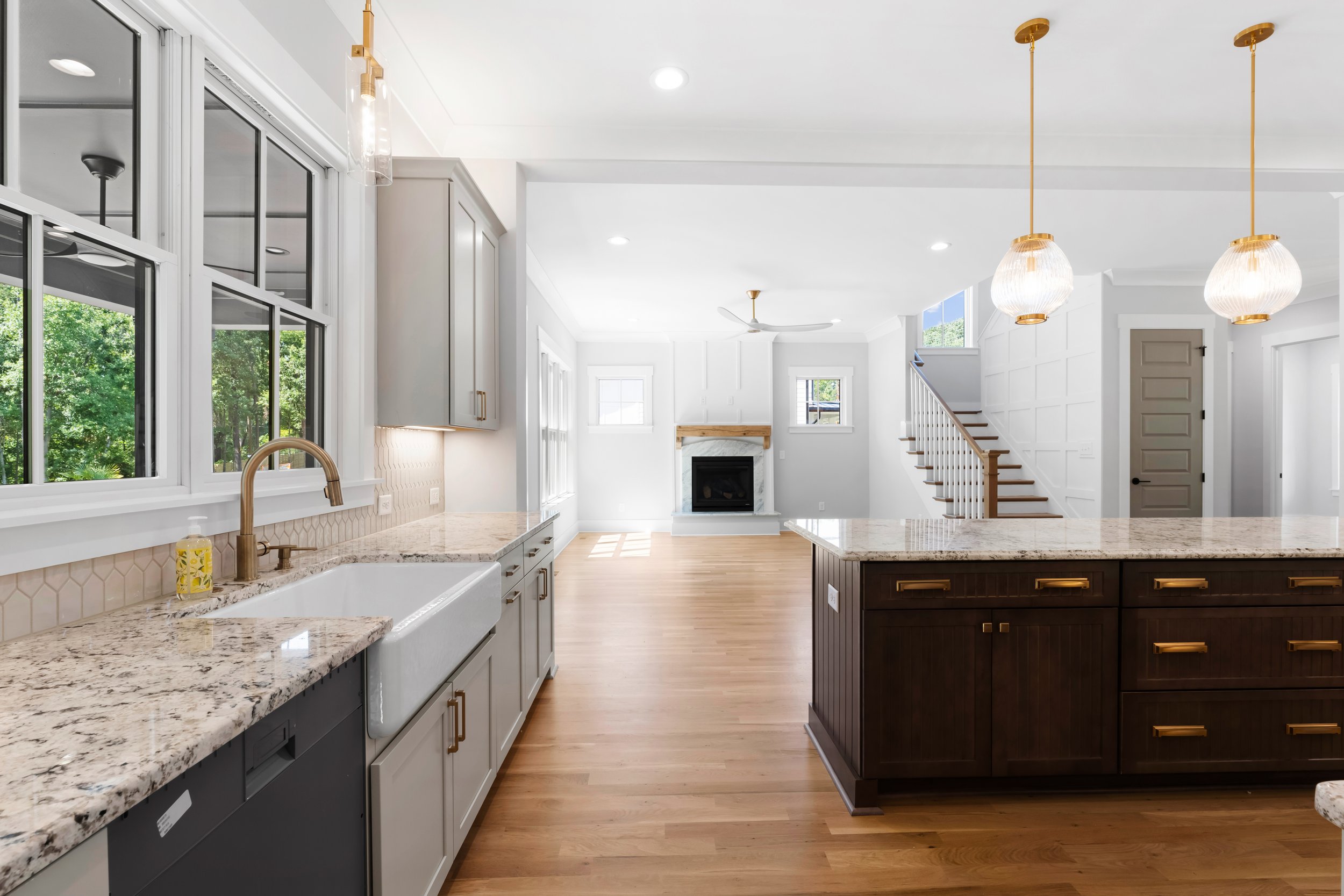 Open-concept kitchen and living room with white walls, hardwood floors, granite countertops, gray and dark wood cabinets, large windows, fireplace, staircase with white spindles, and pendant lights.