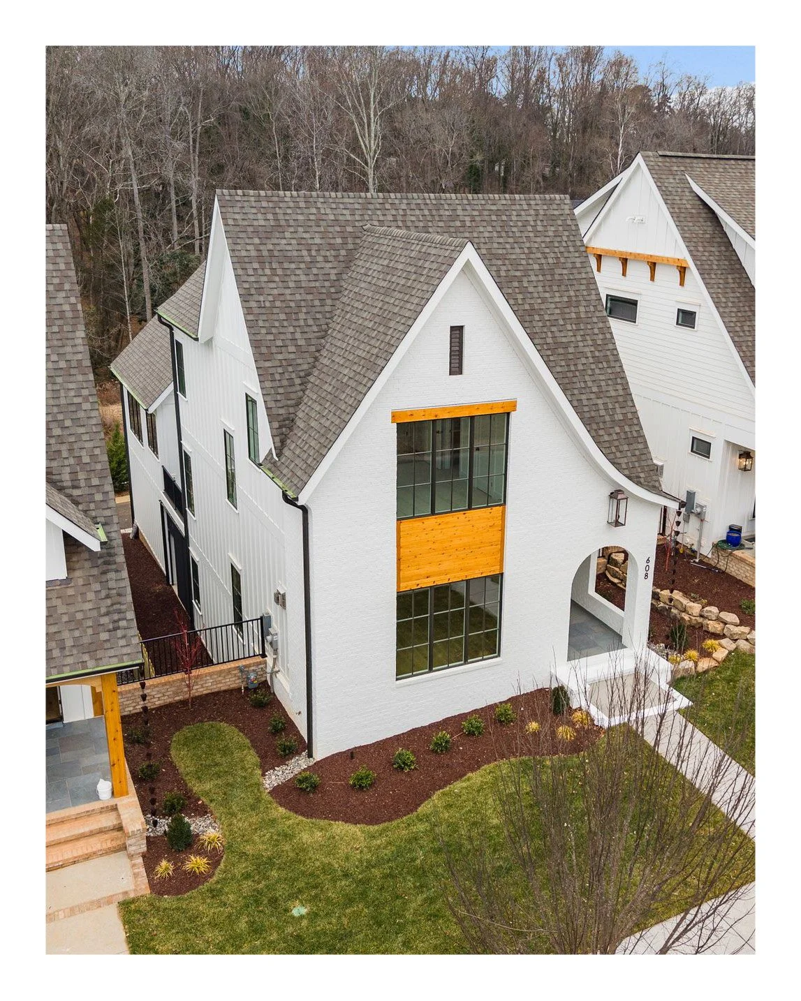A modern white house with large windows and a gabled roof, surrounded by a landscaped yard with grass, small bushes, and trees, on a residential street with other similar houses and wooded area in the background.