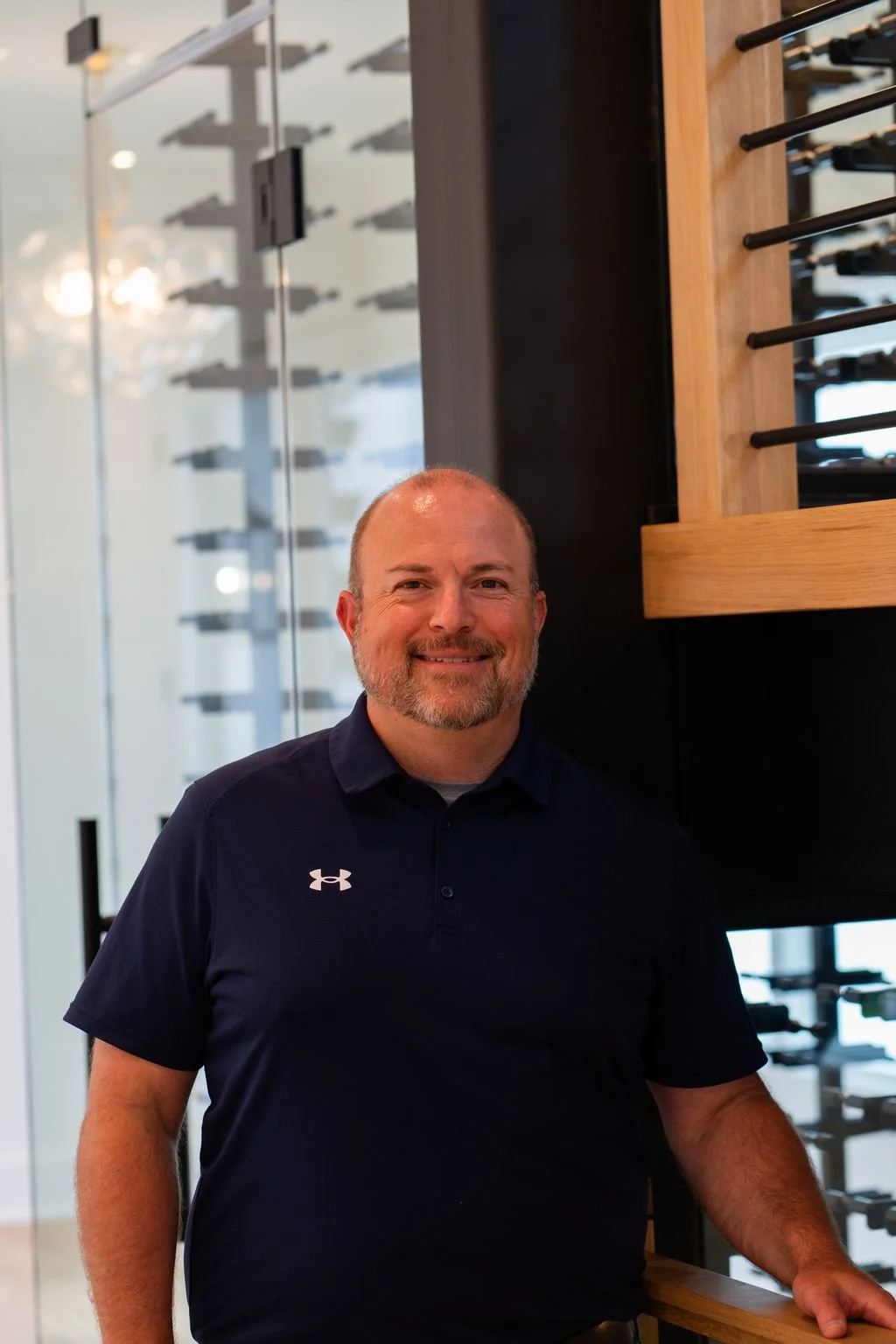 A middle-aged man with a beard smiling, wearing a dark Under Armour polo shirt, standing inside a store with wine racks and glass walls in the background.