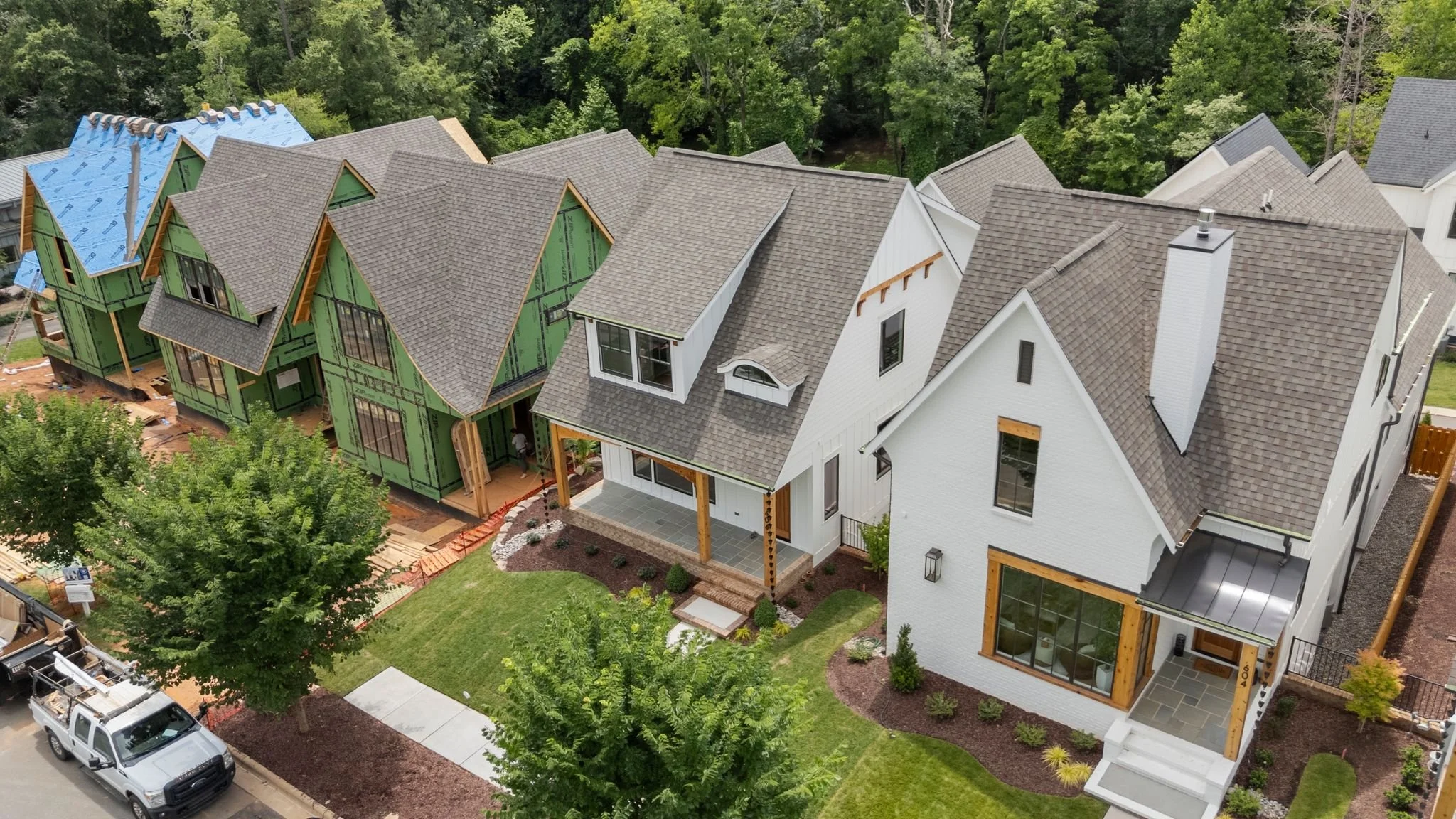 Aerial view of a row of houses under construction, with one house fully built and others in various stages of framing and roofing, surrounded by green trees.