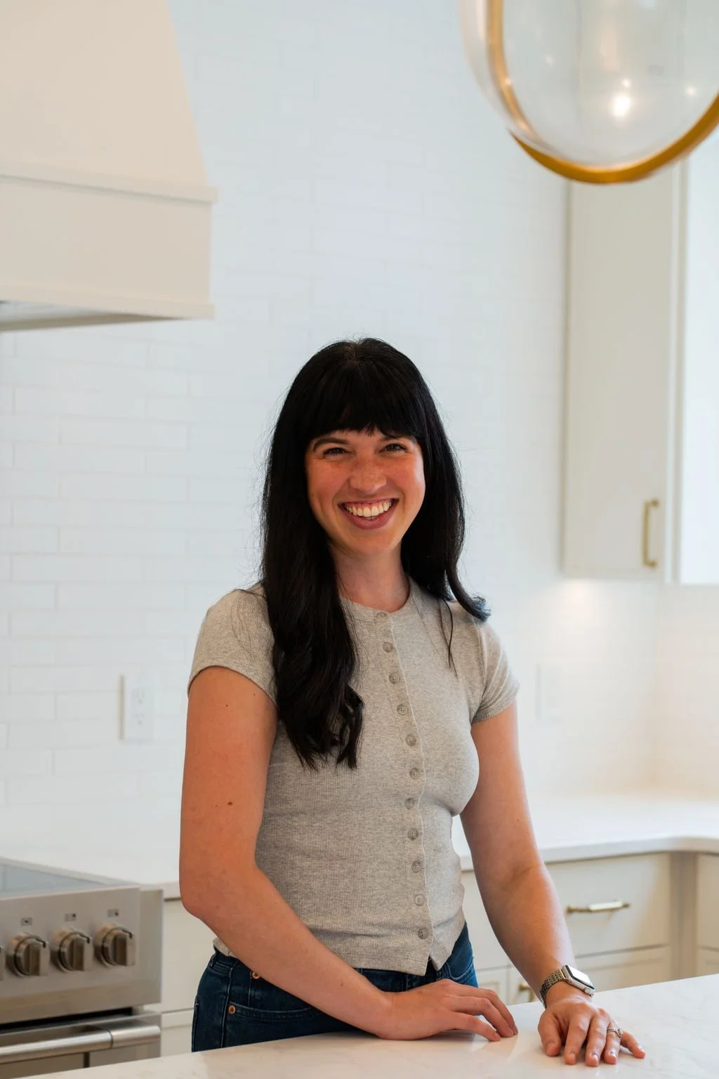 A woman with long dark hair and bangs smiling in a bright white kitchen, wearing a light gray button-up shirt and a watch on her left wrist.