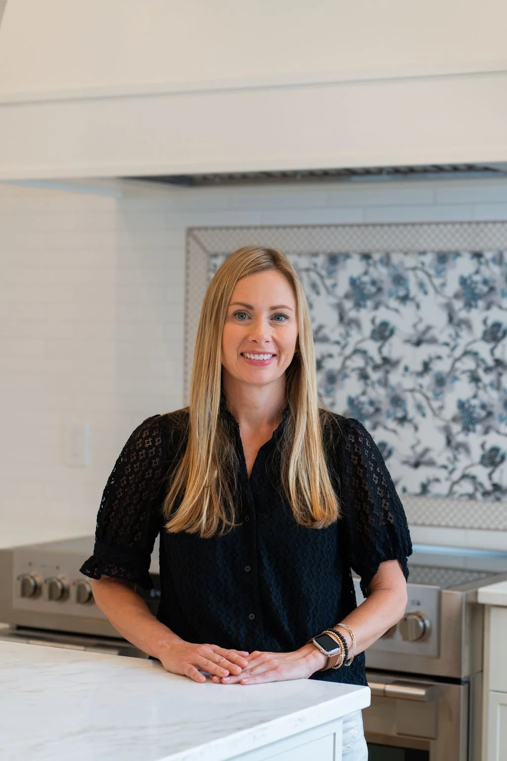 A woman with long blonde hair and a black lace blouse standing in a modern kitchen, smiling, with her arms resting on a white countertop.