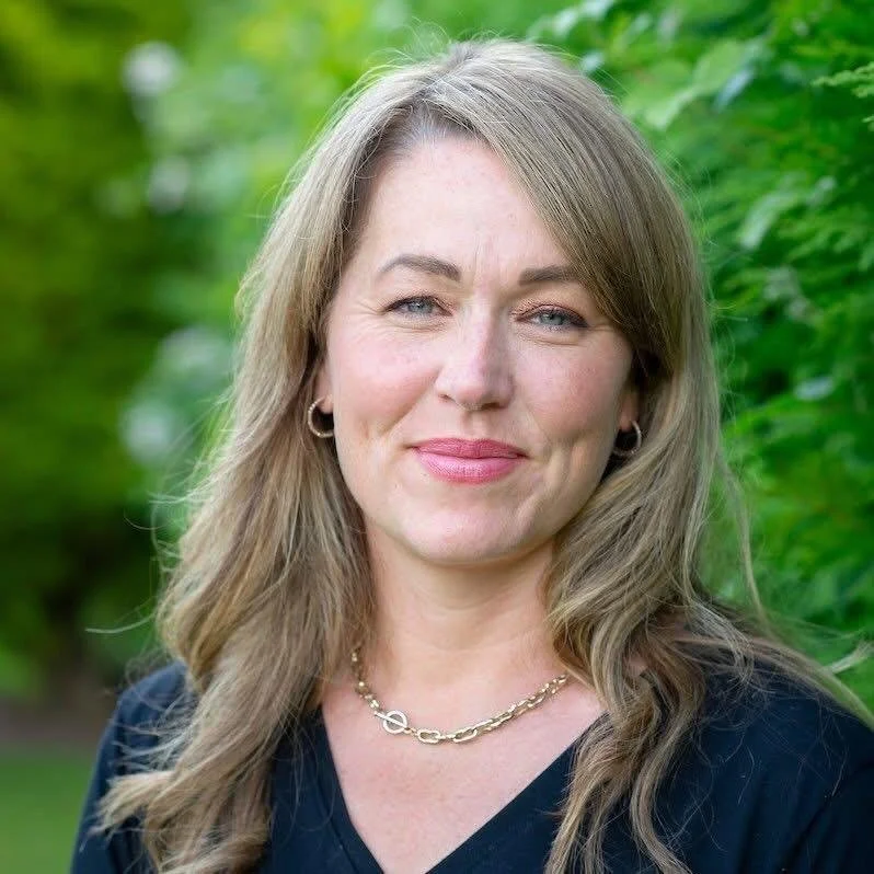 A woman with long light brown hair and blue eyes, smiling slightly, wearing hoop earrings and a gold necklace, outdoors with green foliage in the background.