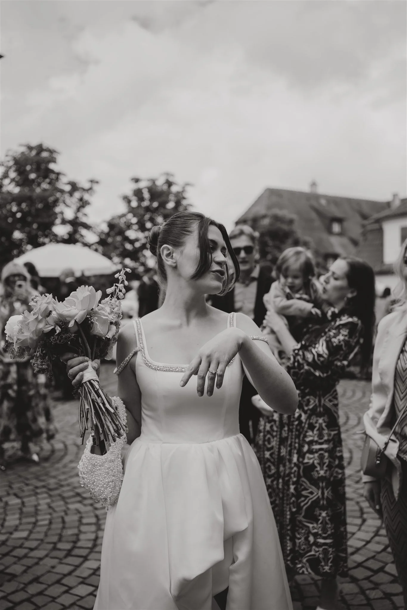 Schwarz-weiß-Foto einer jungen Frau in einem Brautkleid mit einem Blumenstrauß in der Hand, umgeben von mehreren Menschen auf einer Kopfsteinpflasterstraße, vermutlich bei einer Hochzeit.