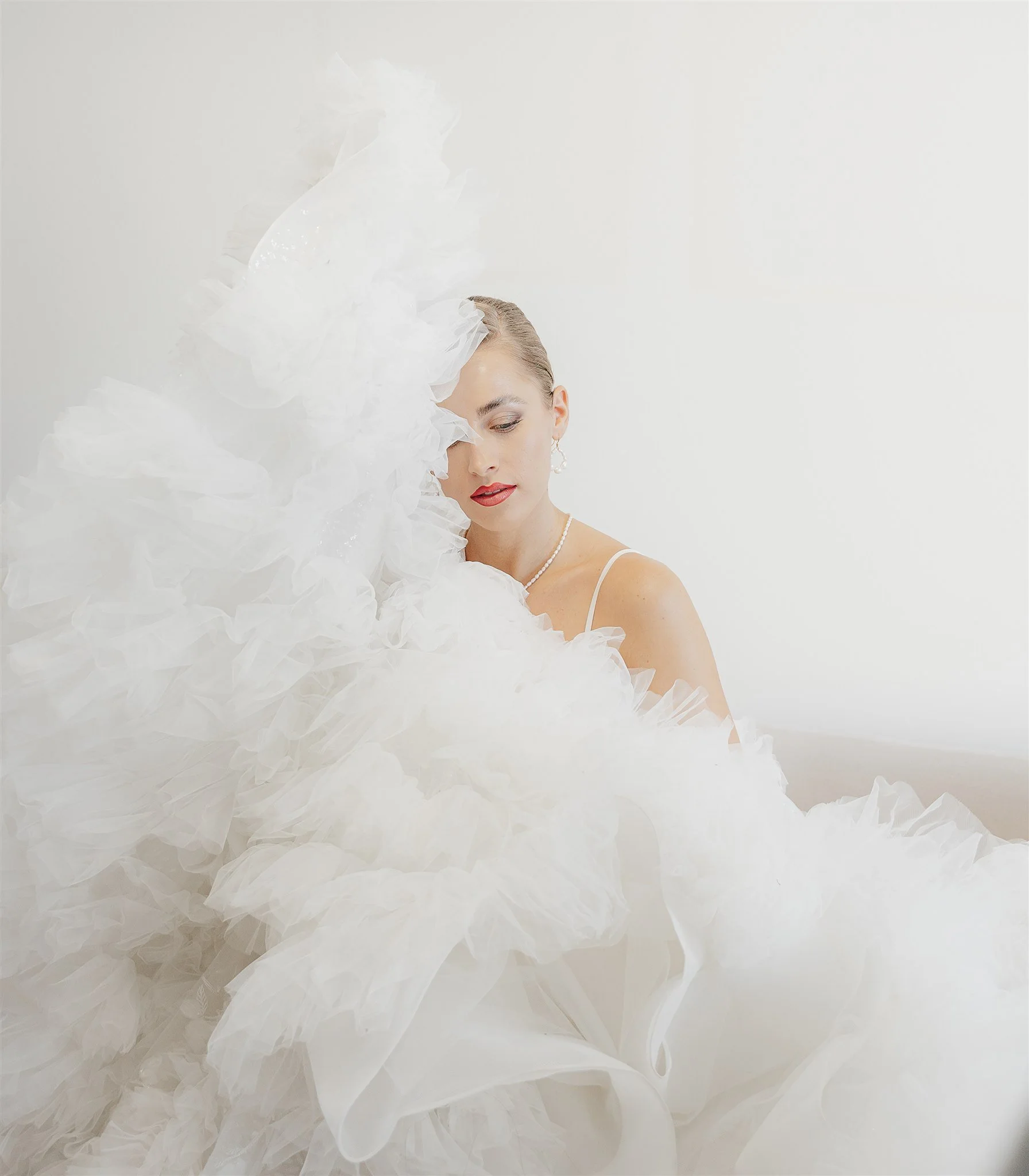 Frau in Hochzeitskleid mit glitzernder Perlenkette und Ohrringen, posiert vor weißem Hintergrund, umgeben von weißen Tüll- und Organza-Falten.