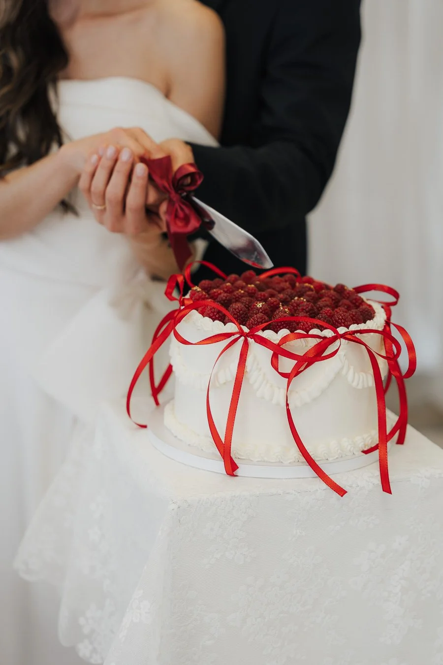 Ein Brautpaar schneidet eine Hochzeitstorte mit roten Beeren und roten Schleifen, auf einem weißen Tischtuch.