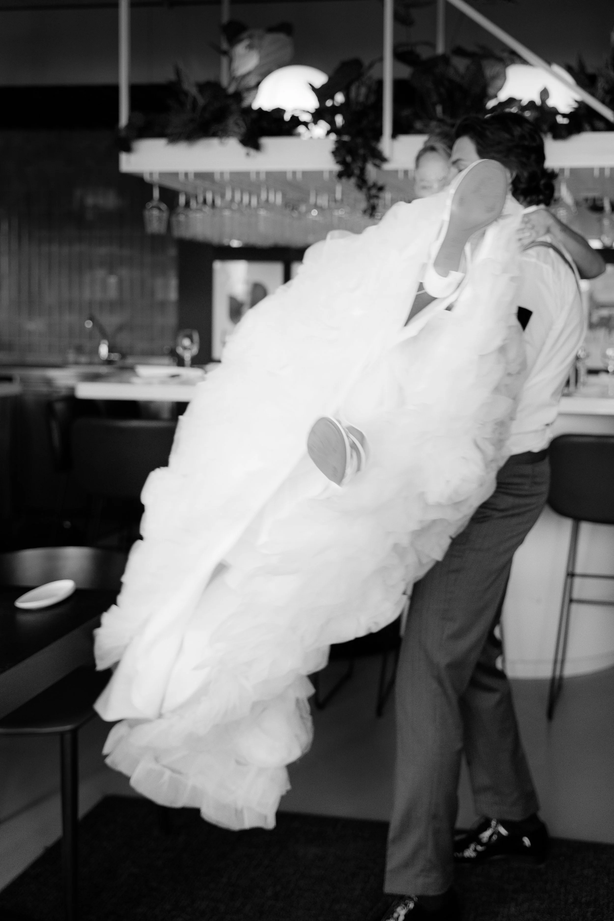 Ein Mann trägt eine sehr große weiße Brautkleid, während sie gemeinsam tanzen oder posieren in einem Restaurant. Das Bild ist schwarz-weiß.