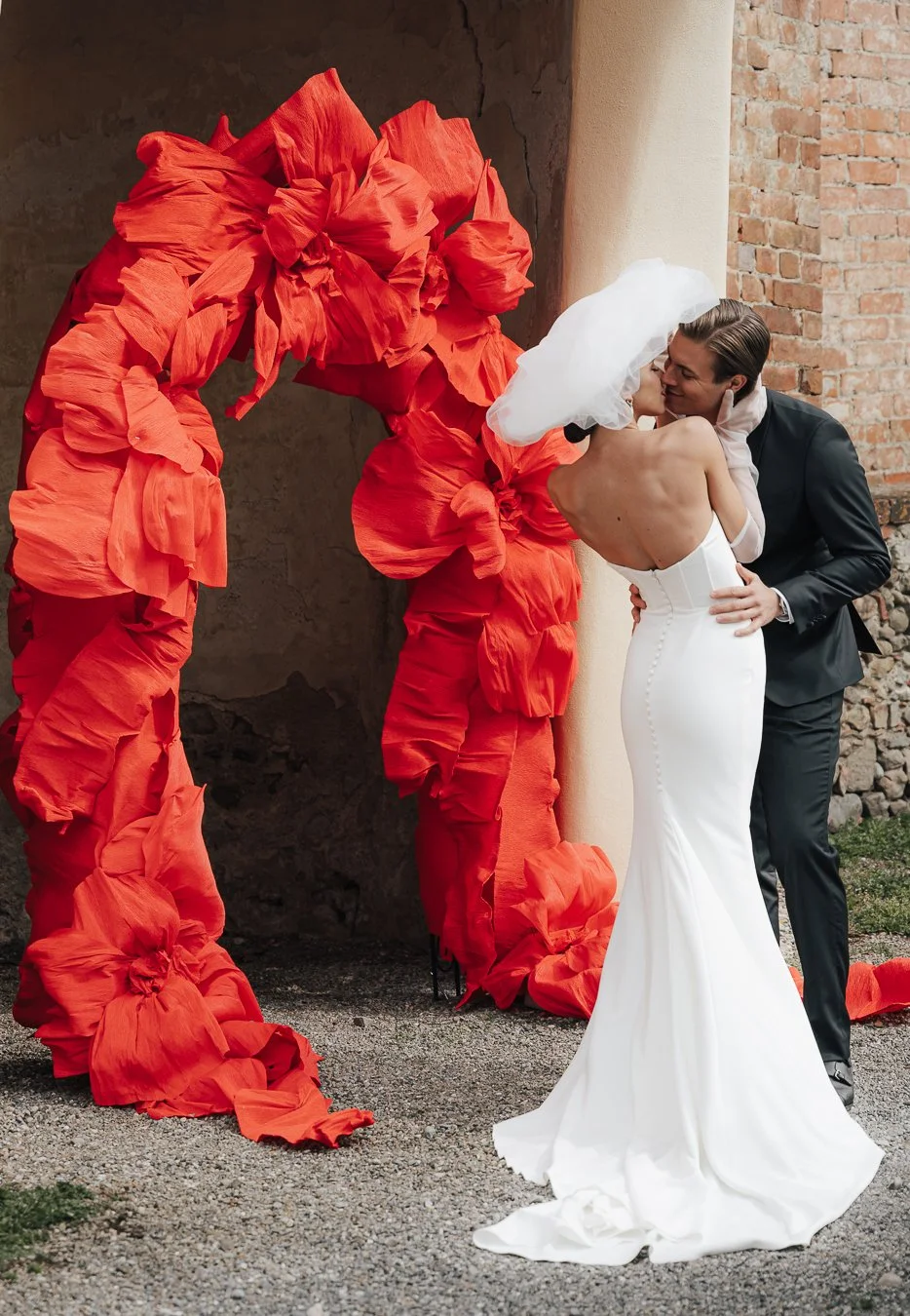 Ein Hochzeitspaar küsst sich vor einer roten, skulpturalen Wand aus Stoff, die an eine Wolke oder Blüten erinnert. Die Braut trägt ein schulterfreies, langes weißes Kleid mit Schleier, der Bräutigam einen schwarzen Anzug.