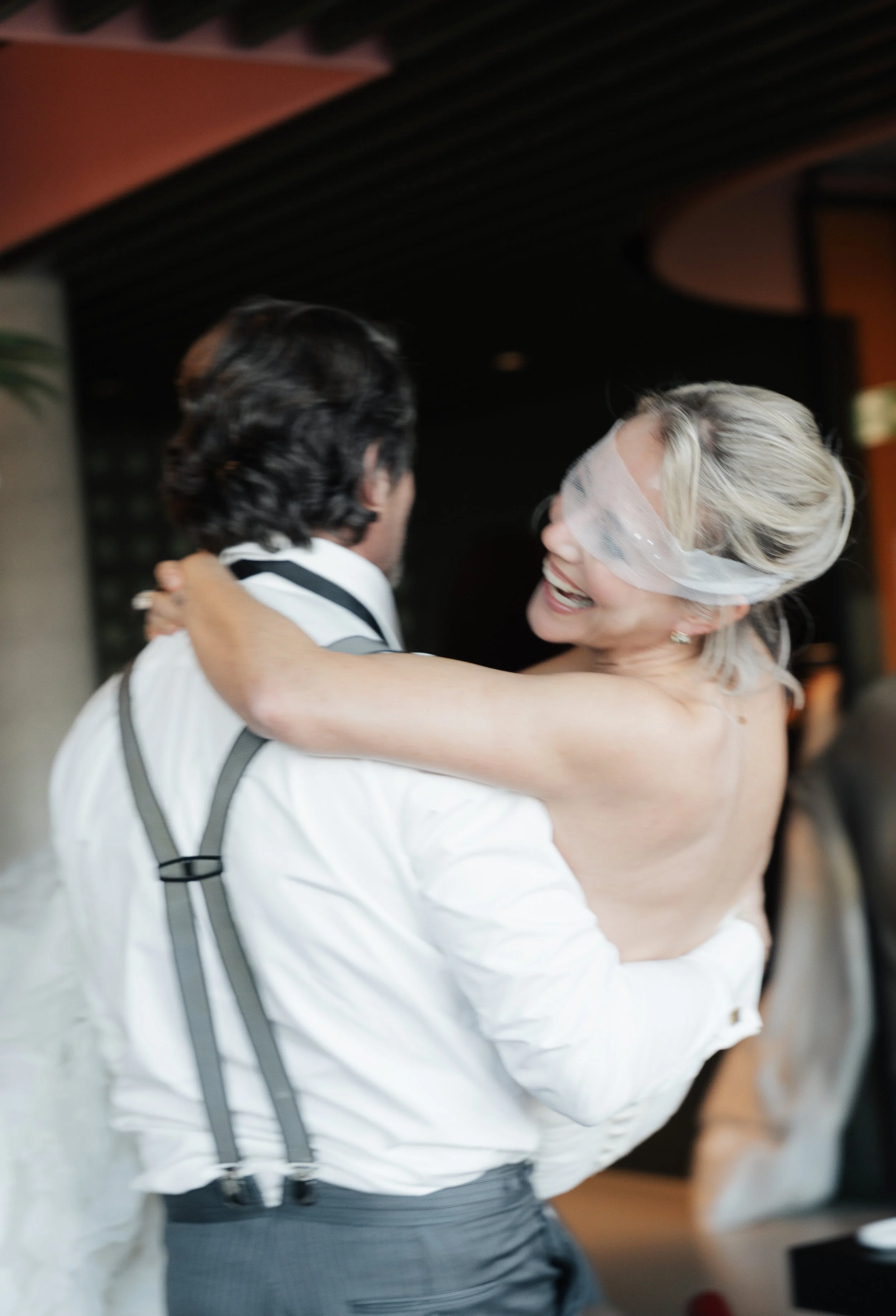 Ein Mann hebt eine lachende Frau in einem weißen Kleid, die ein schützendes Tuch über den Augen trägt, bei einer festlichen Gelegenheit.