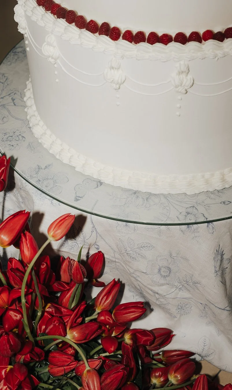 Eine große weiße Geburtstagstorte mit rotem Fruchtbelag auf einem mit Blumenmuster bedruckten Tischtuch, umgeben von roten Tulpen.