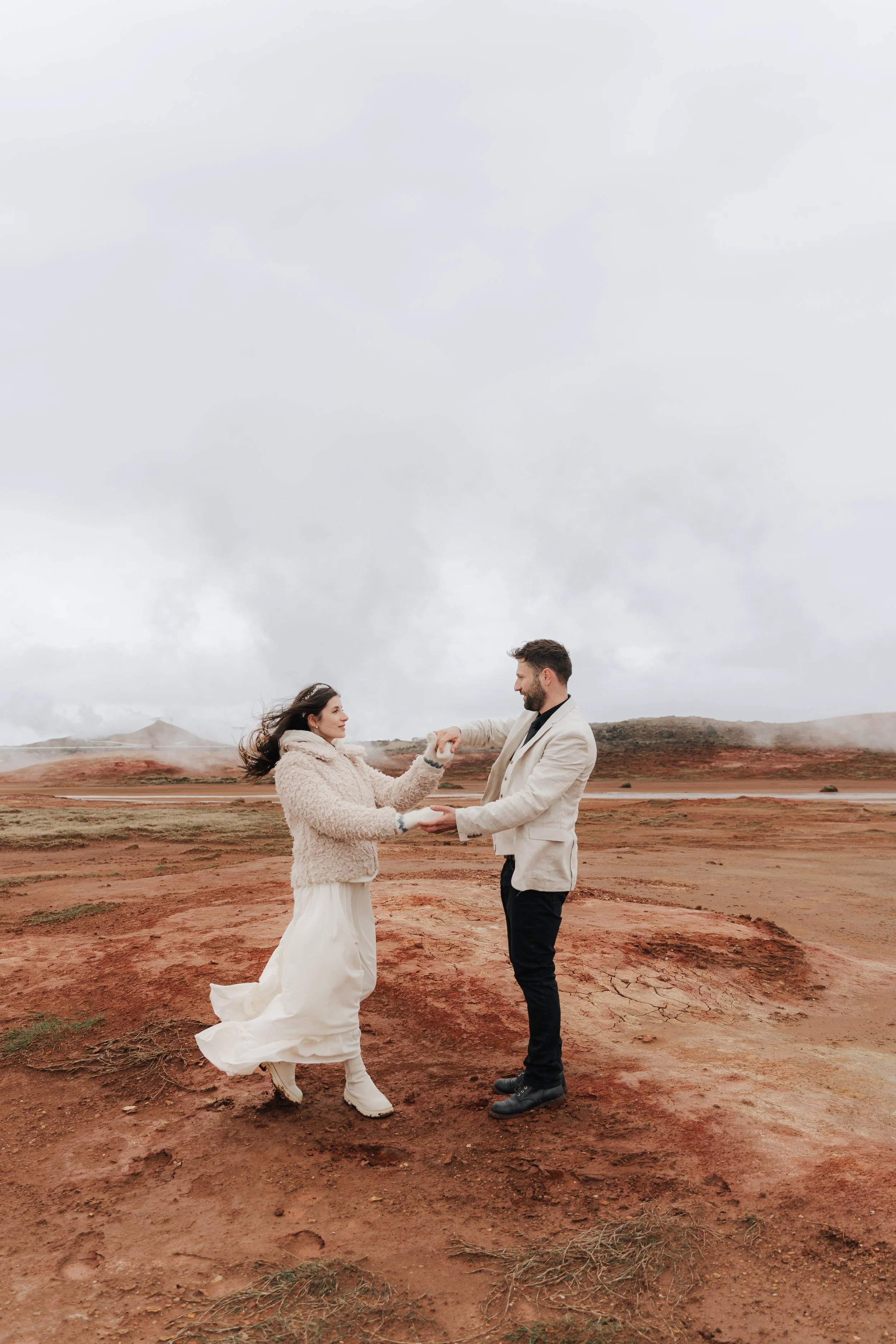 Ein Paar steht in einer kargen, roten Wüstenlandschaft, hält sich an den Händen und sieht sich liebevoll an, während die Wolken den Himmel bedecken.