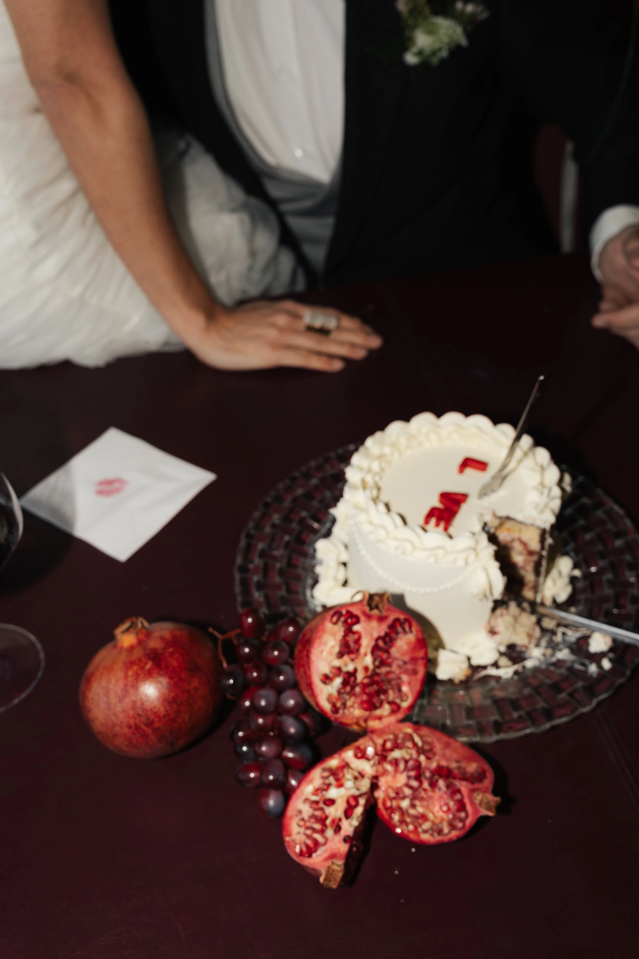 Hochzeitstorte mit weißen Verzierung, umgeben von Granatäpfeln und Weintrauben auf einem dunklen Tisch.