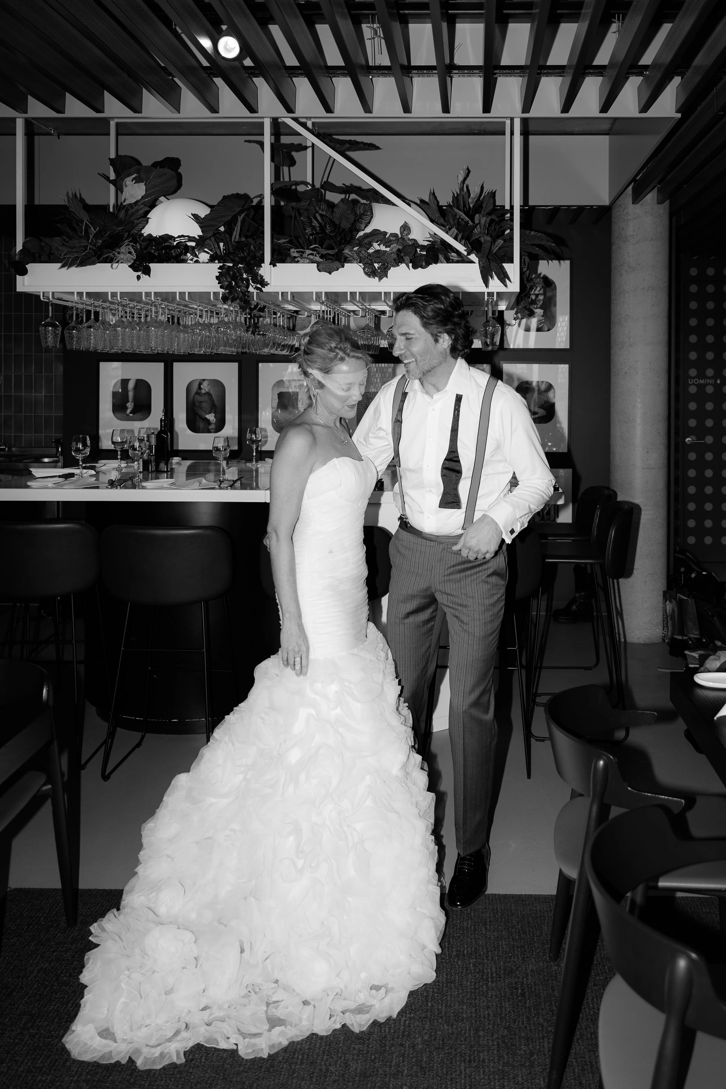 Ein Brautpaar beim Tanzen in einem Restaurant, die Braut trägt ein langes, rosenverziertes Hochzeitskleid, der Bräutigam trägt eine Hose mit Hosenträgern. Im Hintergrund sind Wein- und Gläser auf dem Bar-Theke zu sehen.