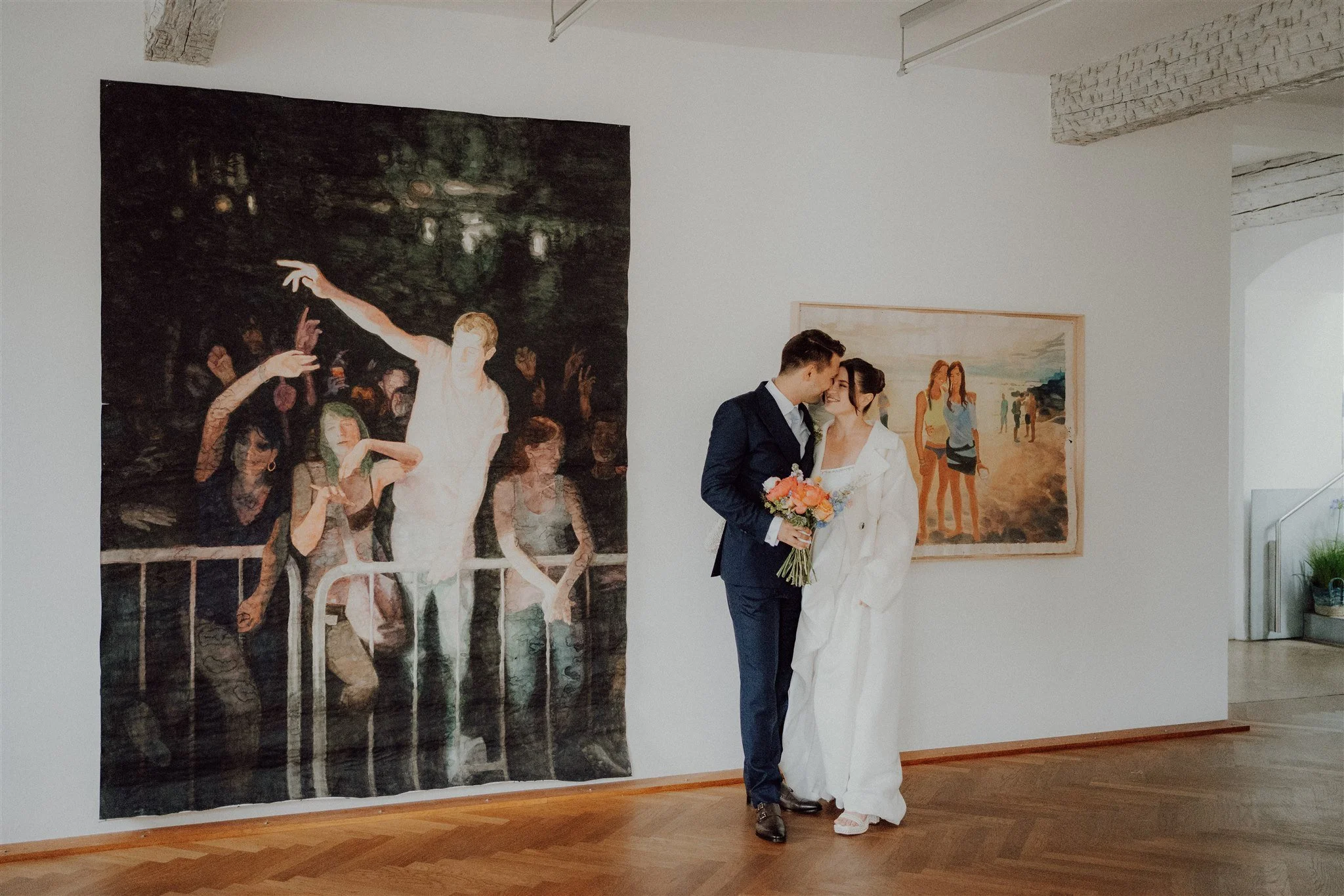 Ein Paar, vermutlich Braut und Bräutigam, steht in einem Kunstmuseum vor zwei Gemälden und sieht sich liebevoll an. Die Braut hält einen Blumenstrauß.