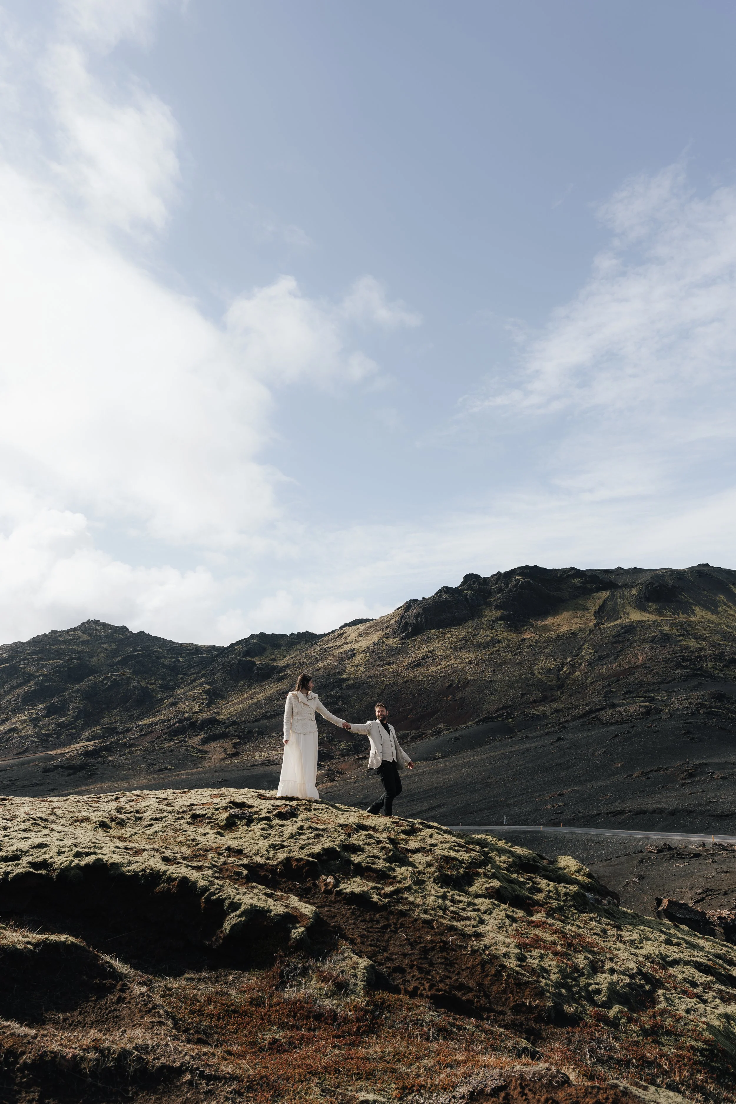 Ein Paar in formeller Kleidung hält sich an einem felsigen, grünen Hang in einer bergigen Landschaft mit Himmel im Hintergrund.