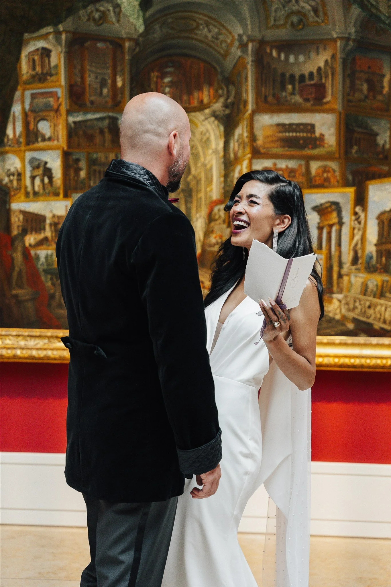 Ein Mann und eine Frau lachen in einer Kunstgalerie, während sie miteinander sprechen. Im Hintergrund sind zahlreiche Gemälde in goldenen Rahmen zu sehen.