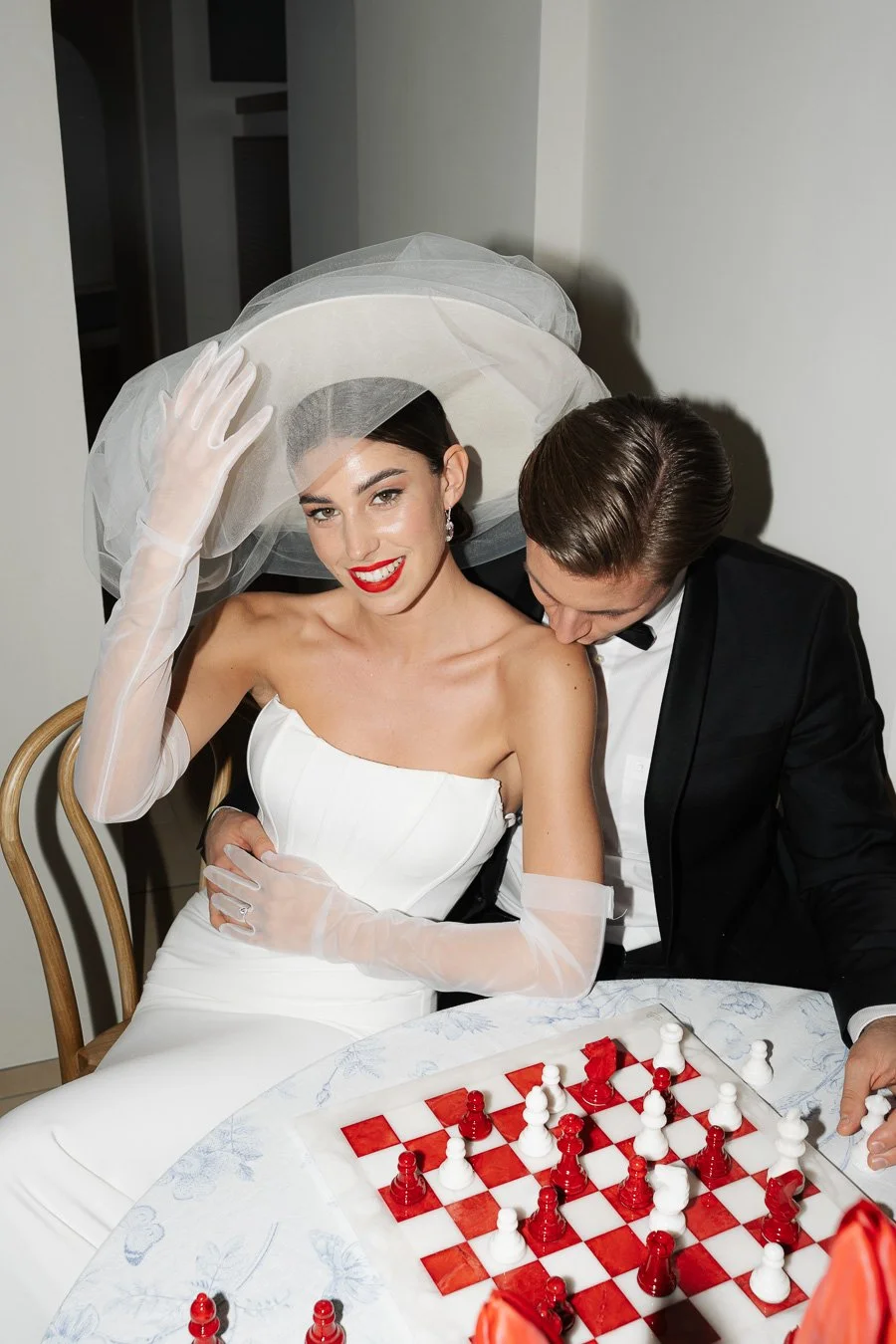 Ein lächelndes Brautpaar spielt Schach, die Braut trägt ein weißes Kleid und einen Schleier, der Bräutigam trägt einen schwarzen Anzug, sie sitzen an einem Tisch mit einem Schachbrett, im Hintergrund eine helle Wand.