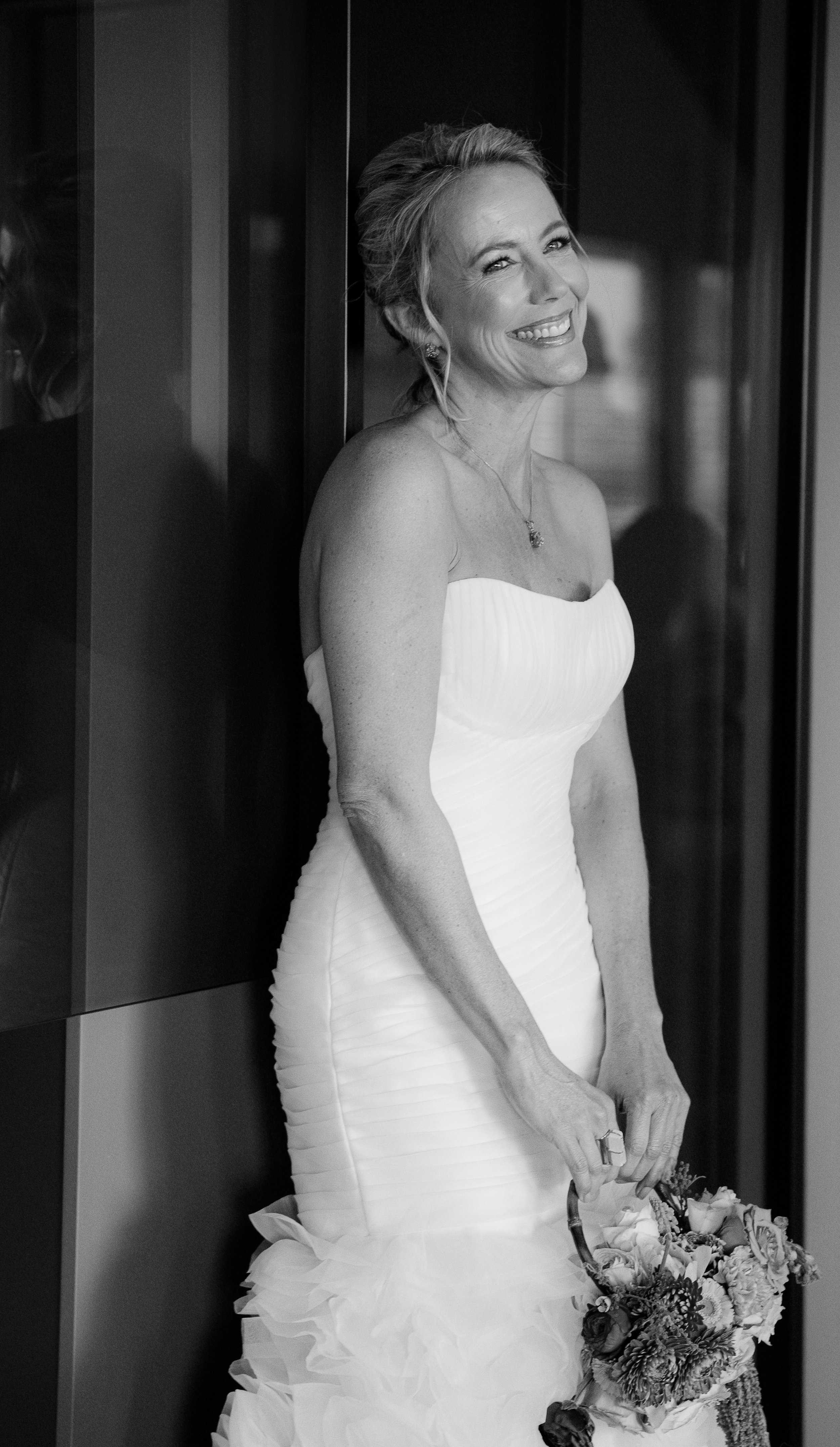 Junge Frau in Hochzeitskleid hält Blumenstrauß, lächelt, steht an dunkler Wand, schwarz-weiß