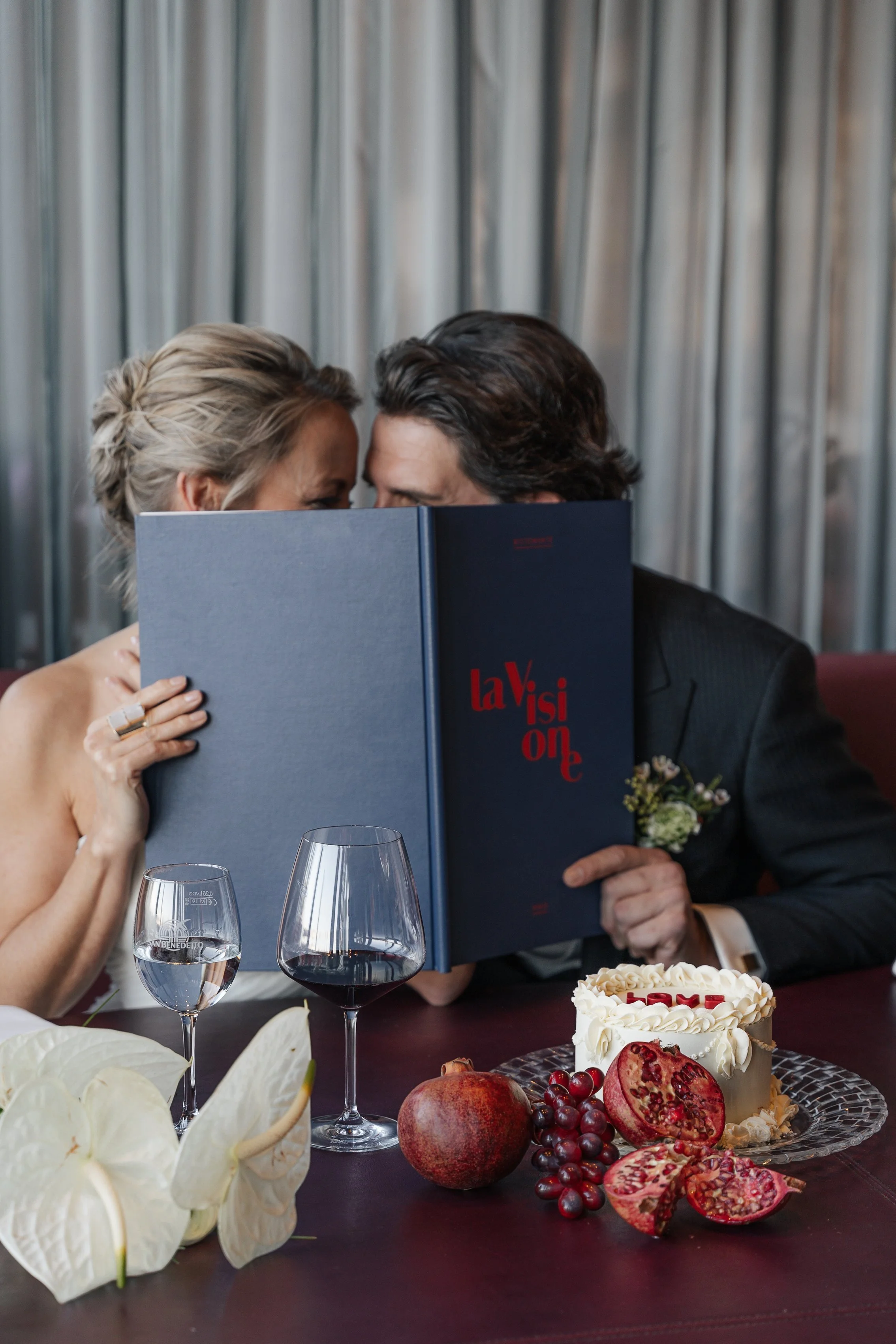 Ein Paar bei einer Hochzeit, das sich hinter einem Buch versteckt, auf einem Tisch mit Wein, Pomegranate, Trauben, einem Hochzeitstorte und Blumen.