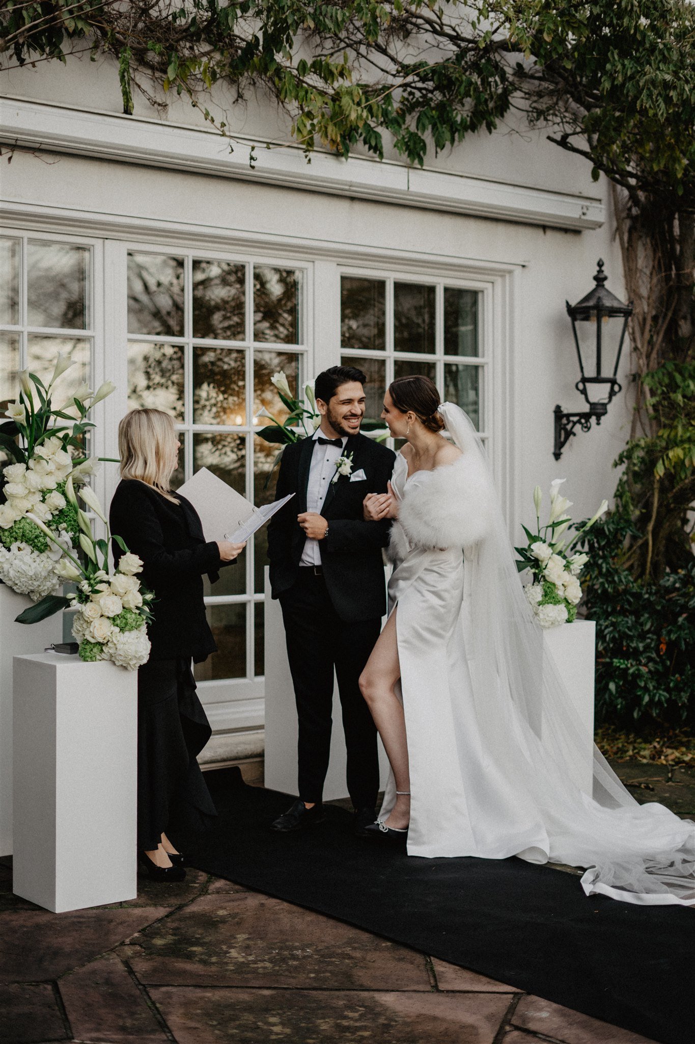 Ein Brautpaar bei der Trauung, der Bräutigam trägt einen schwarzen Anzug und eine Fliege, die Braut trägt ein weißes Hochzeitskleid mit einem langen Schleier und Pelzstola, sie lachen und schauen sich an, eine Zeremonienleiterin liest aus einer Unter