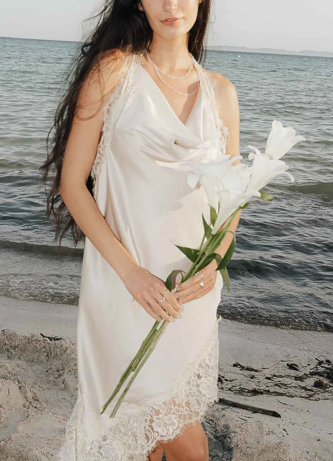 Woman in white linen holding lilies by the sea – Mallorca editorial by Anna van der Veen
