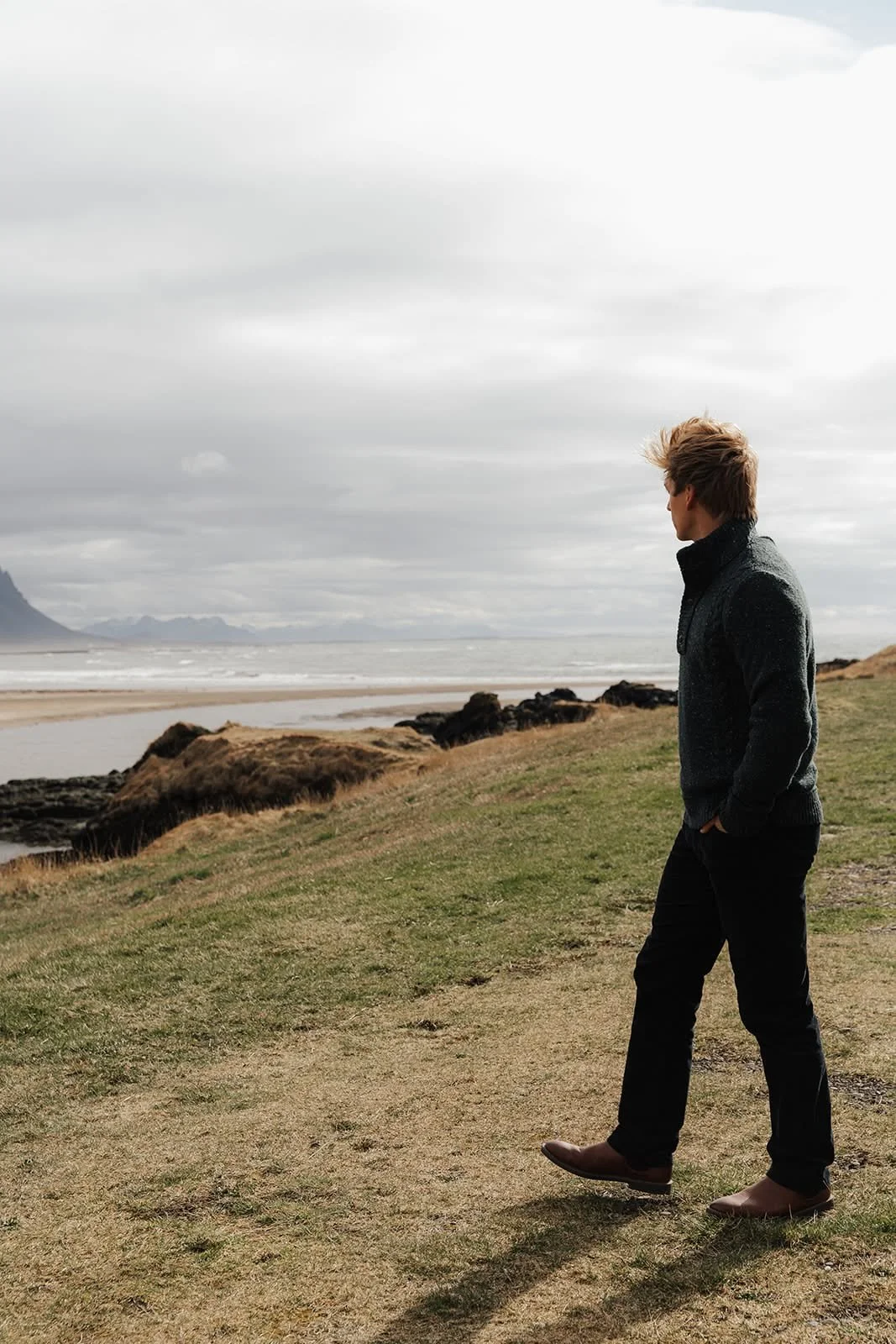 Man in a dark coat walking along the windswept grassy coast near Hótel Búðir, Iceland