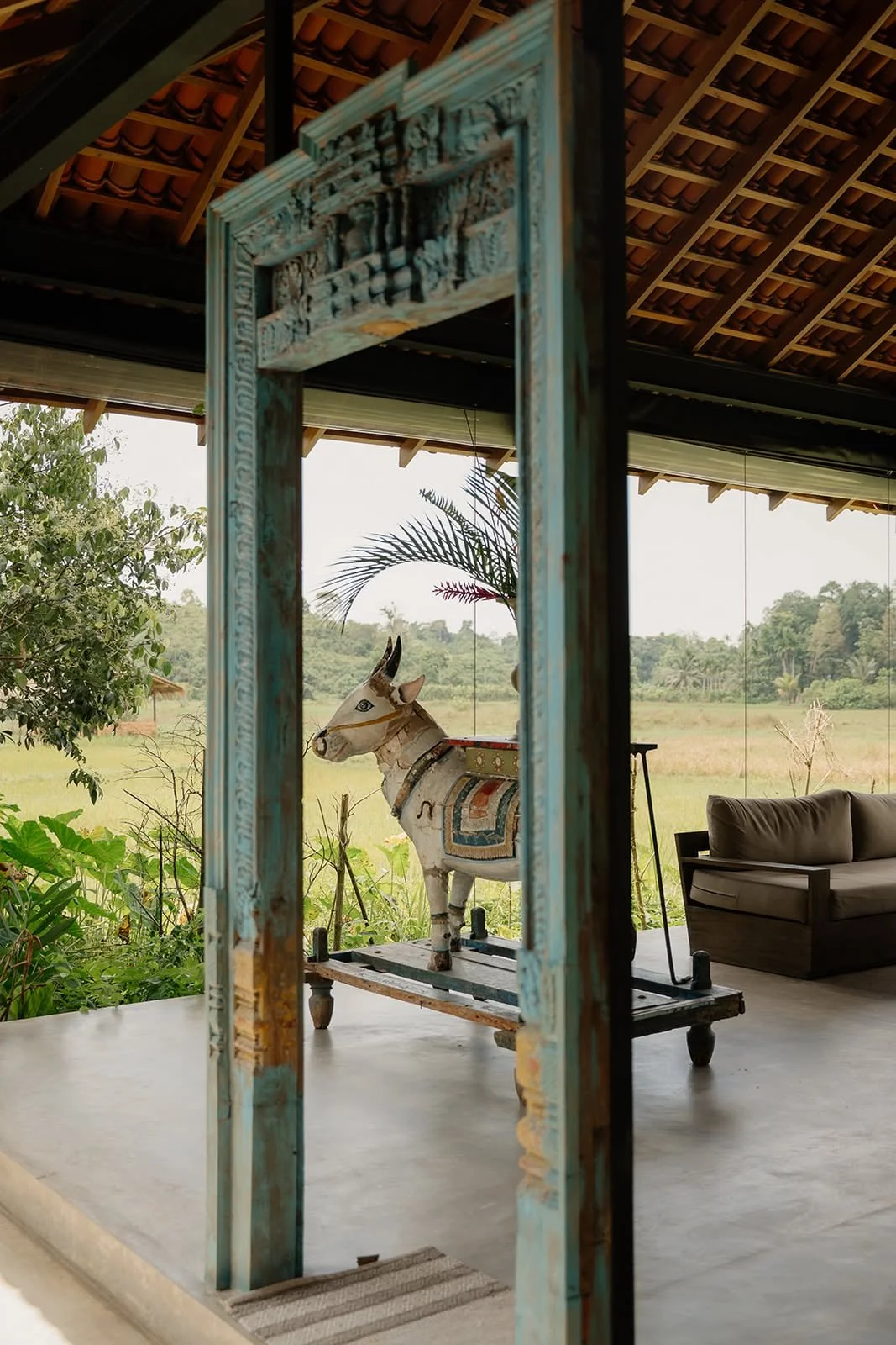 Weathered carved wooden doorway framing rice fields and a deer sculpture at Huma Terra Lodge, Sri Lanka