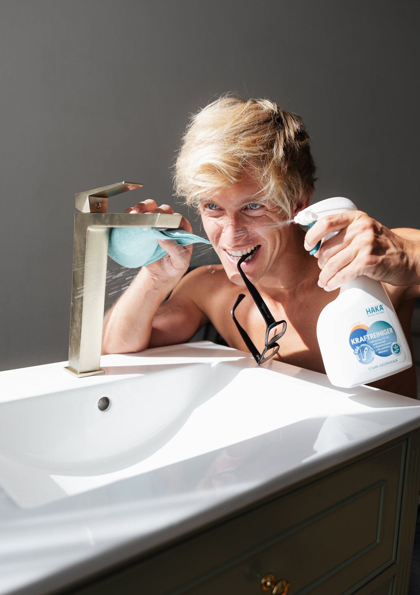 Grinning model spraying HAKA Kraftreiniger on a brass bathroom tap in sunlight