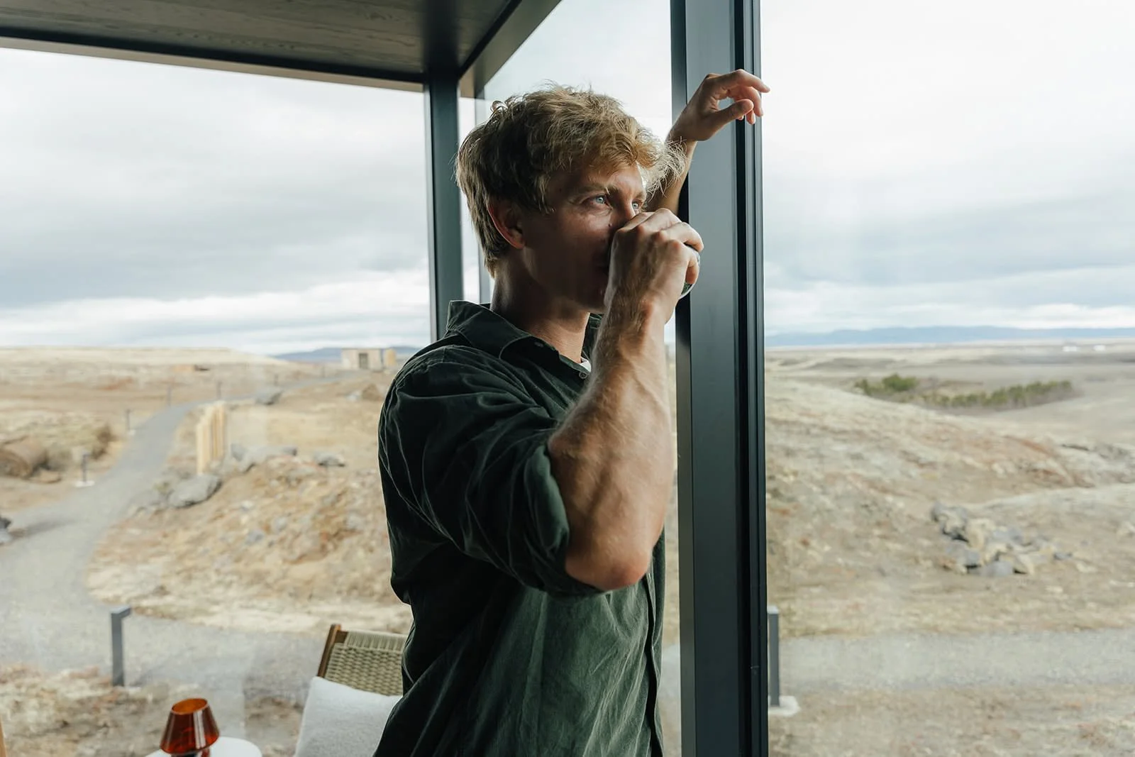 Man drinking coffee at the floor-to-ceiling window of a glass suite at ÖÖD Hekla Horizon, looking out over the Icelandic plain