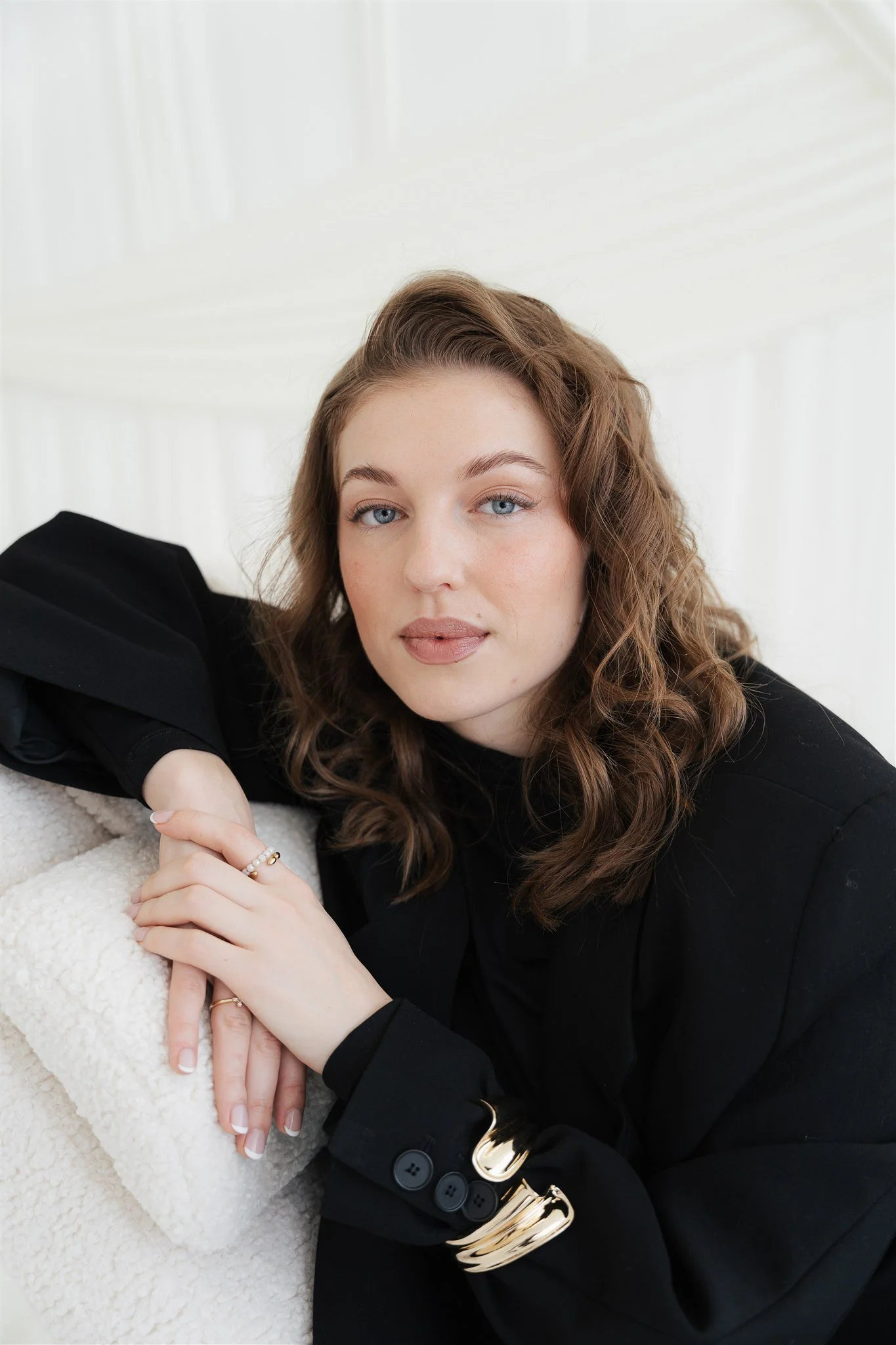 A woman with wavy brown hair, blue eyes, and neutral makeup poses against a light background, wearing a black blazer and jewelry.