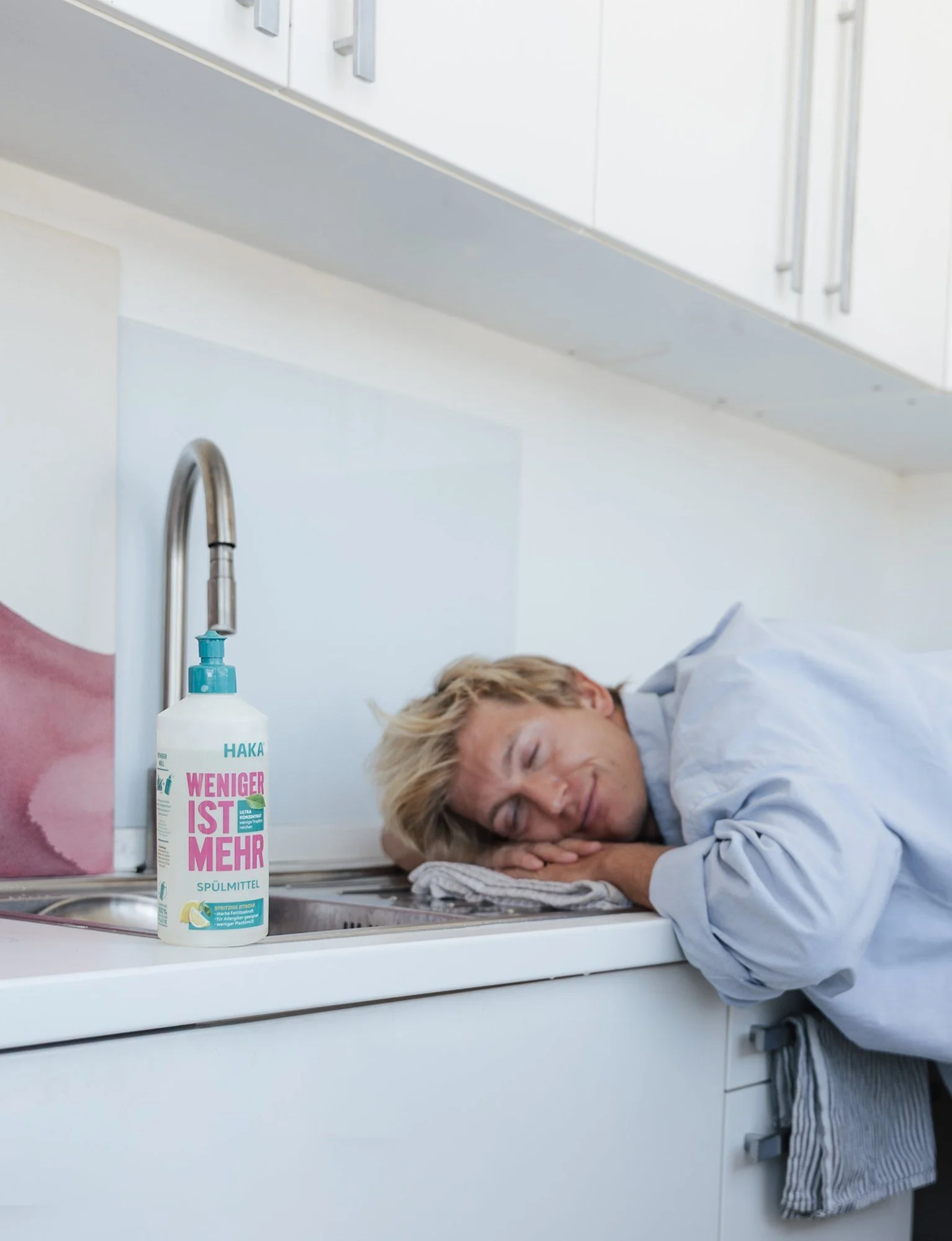 Model napping on a kitchen counter next to a bottle of HAKA dish soap