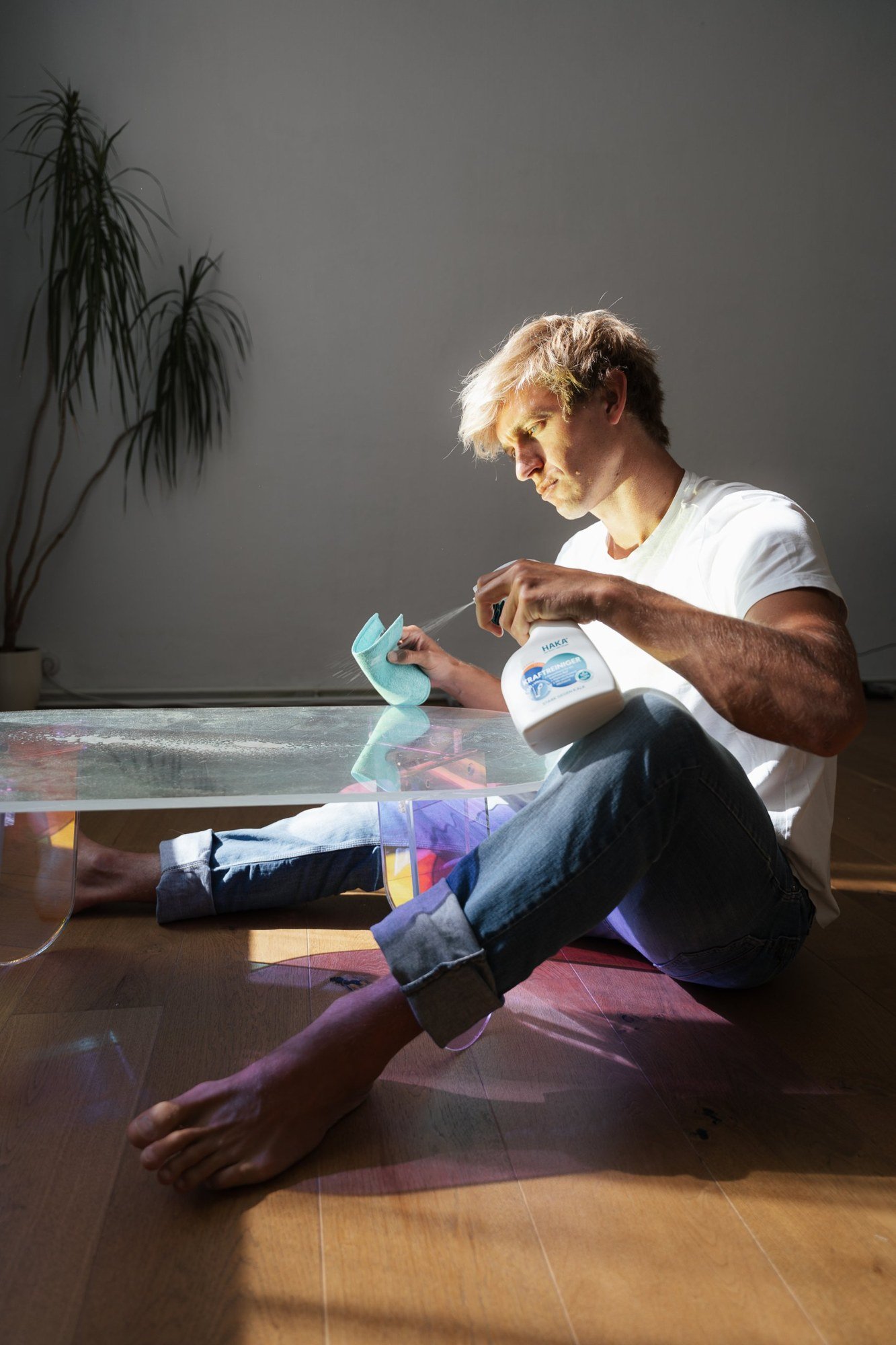 Model sitting on a wooden floor cleaning an iridescent glass table with HAKA spray in sunlight
