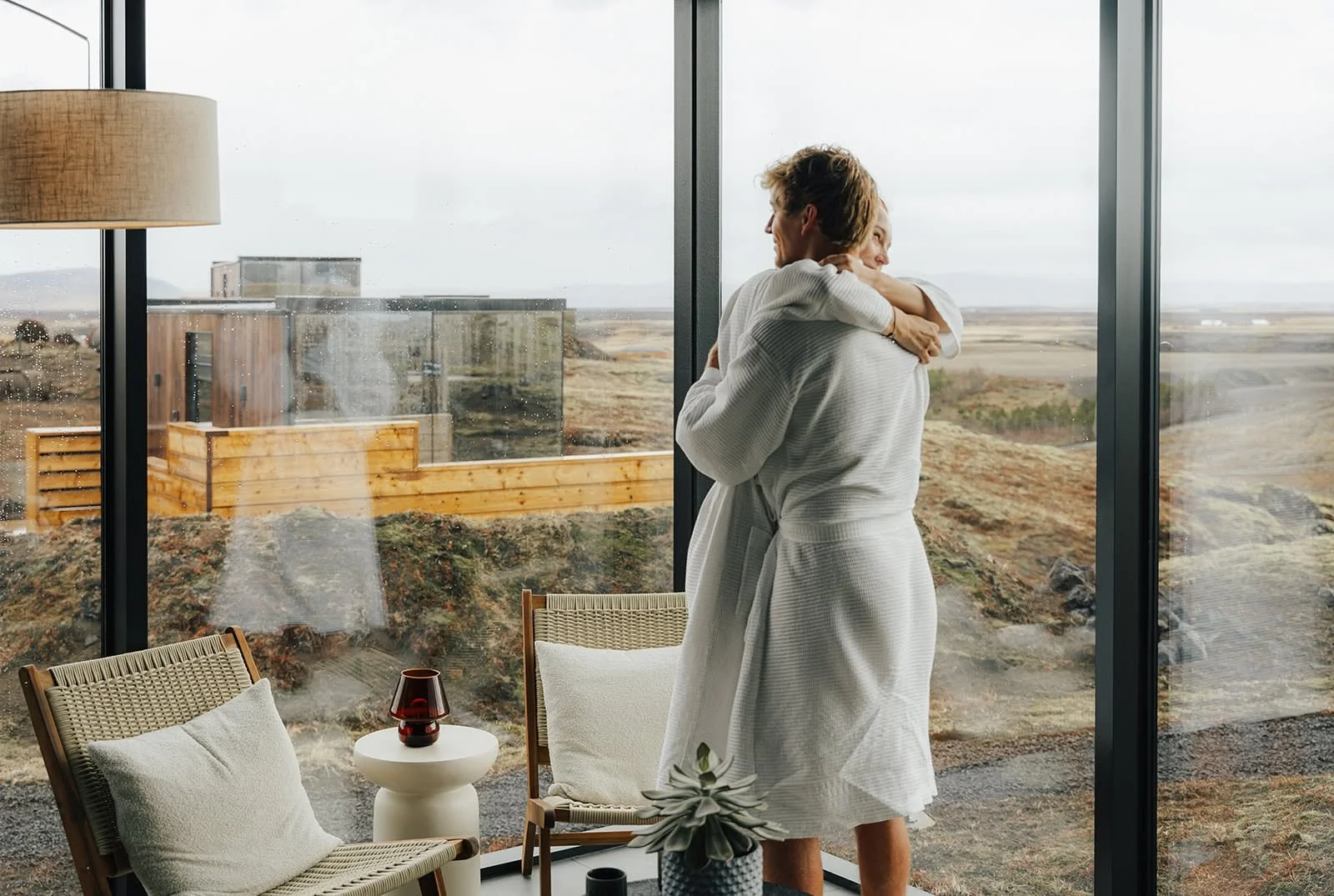 Couple in white bathrobes embracing at the glass wall of an ÖÖD Hekla Horizon mirror suite, with the Icelandic highlands outside