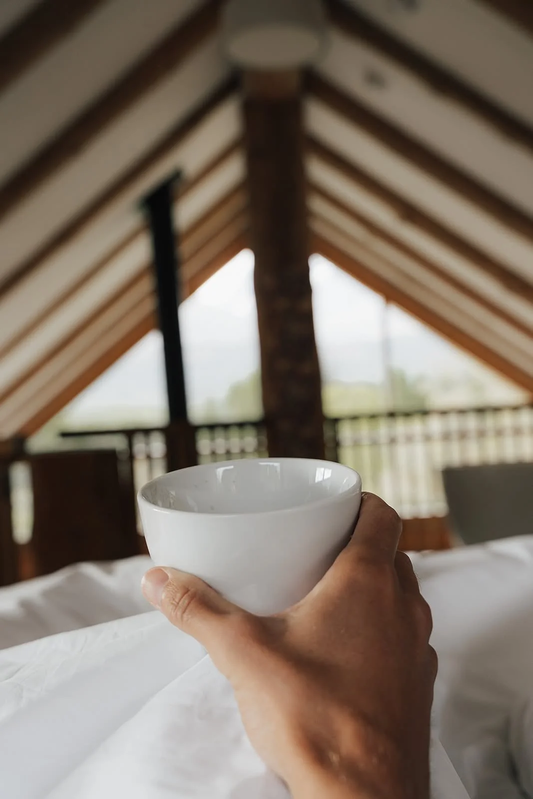 Hand holding a white cup in bed beneath wooden roof beams during a WILD Nurture retreat, Scotland