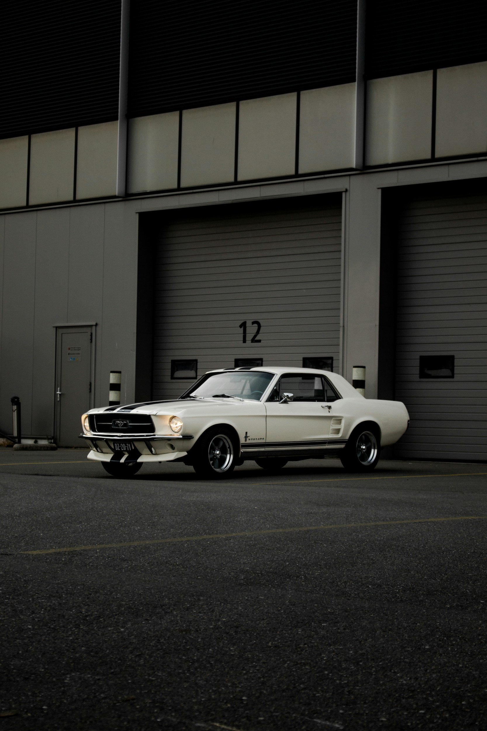 A white classic Ford Mustang parked in front of a large industrial building with a gray roll-up door and black numbered marking 12.