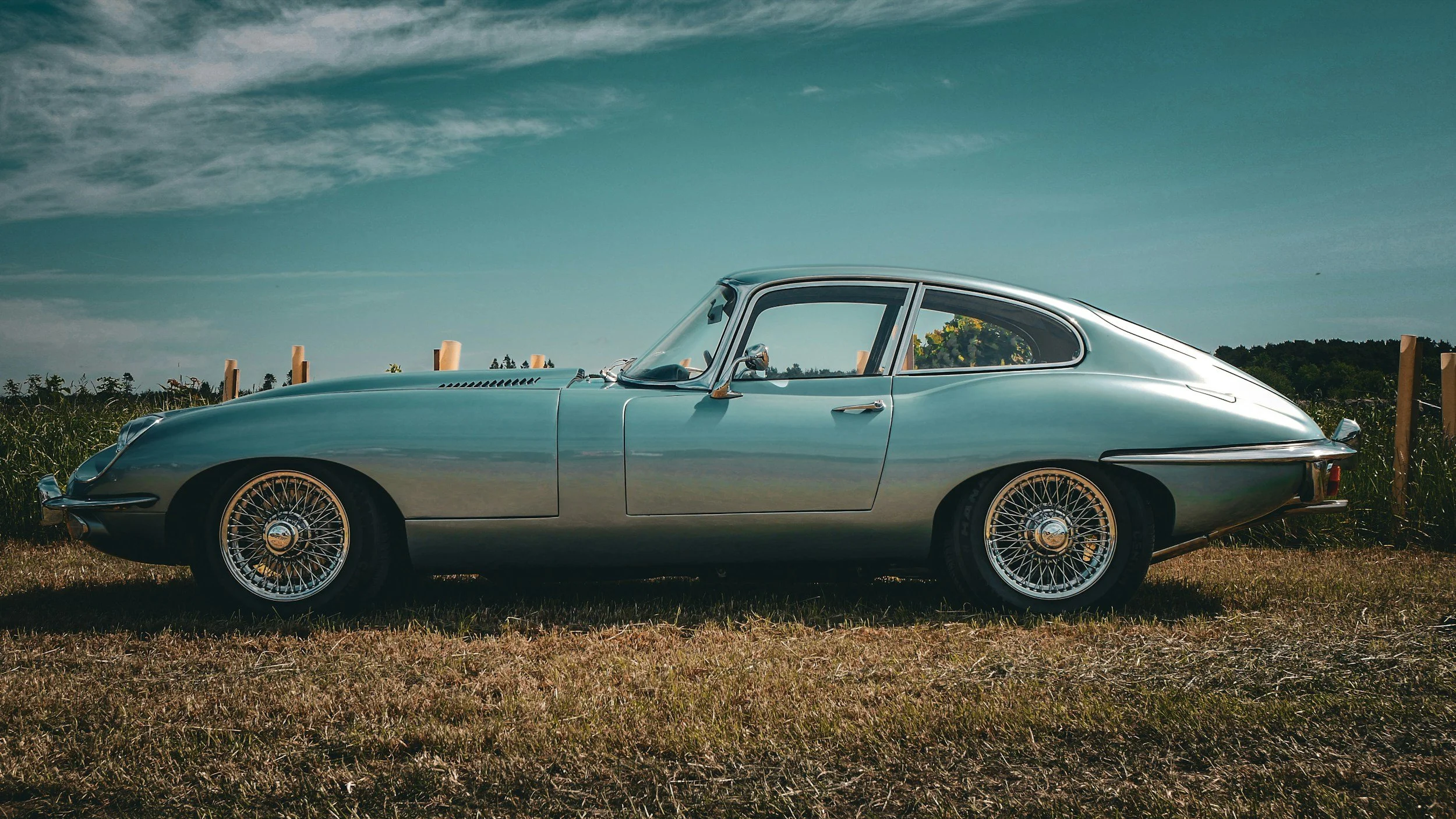 A vintage silver sports car parked on a grassy field with a cloudy sky overhead.