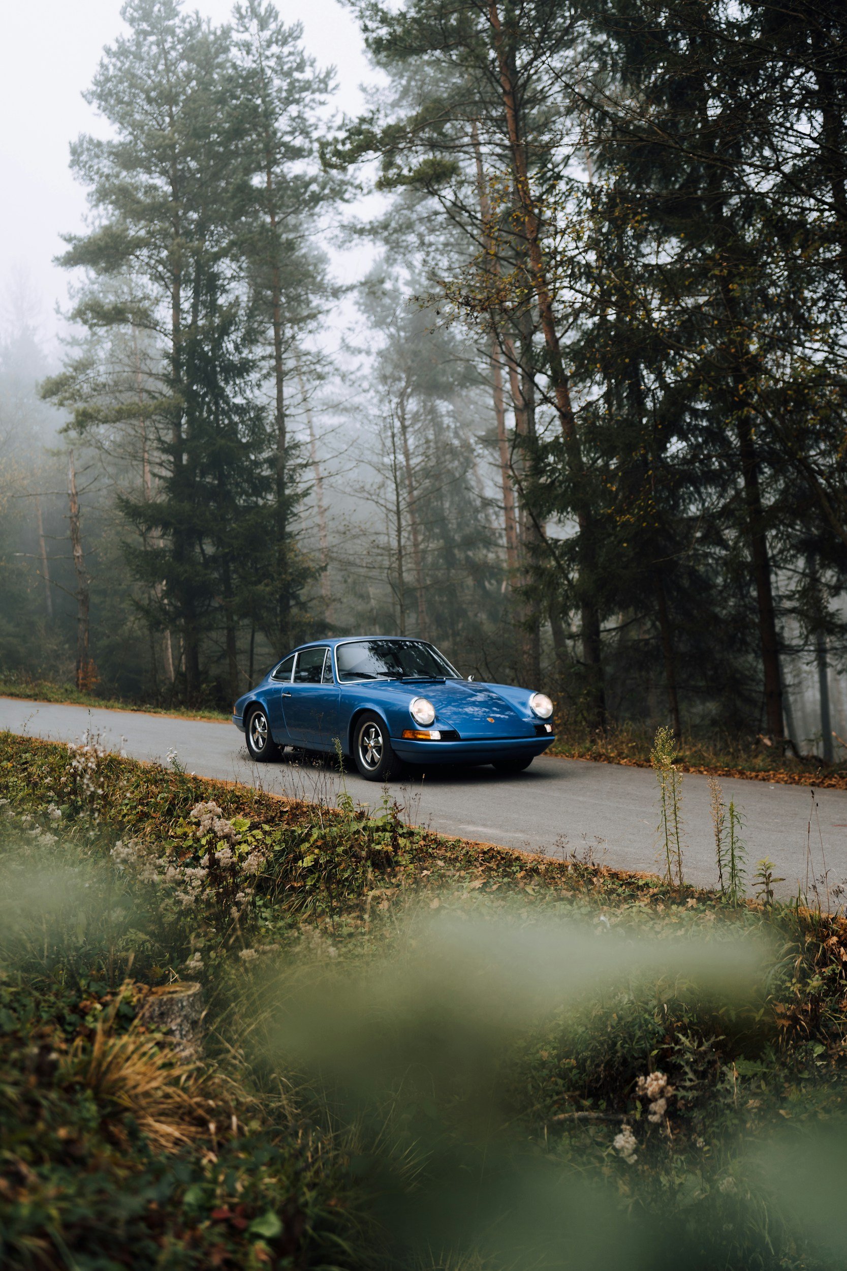 A vintage blue Porsche 911 driving on a forested mountain road with fog in the background.