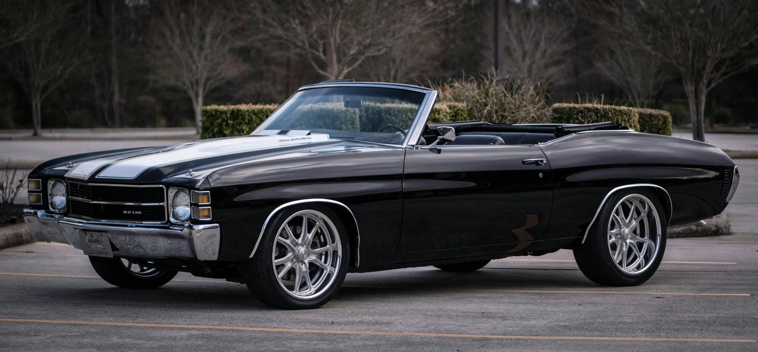 A black vintage convertible car parked in an empty parking lot with trees and bushes in the background.