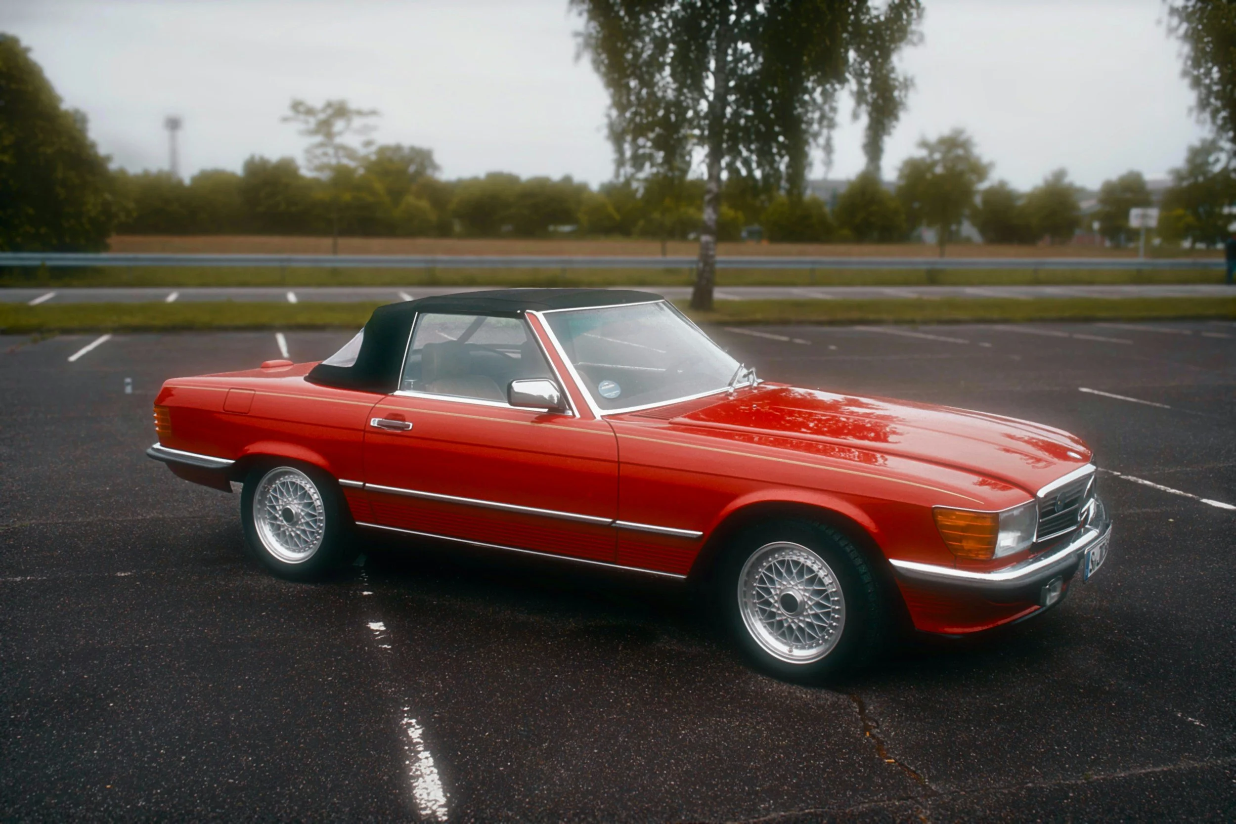 A red classic Mercedes-Benz convertible with a black soft top parked in an empty parking lot on a cloudy day.