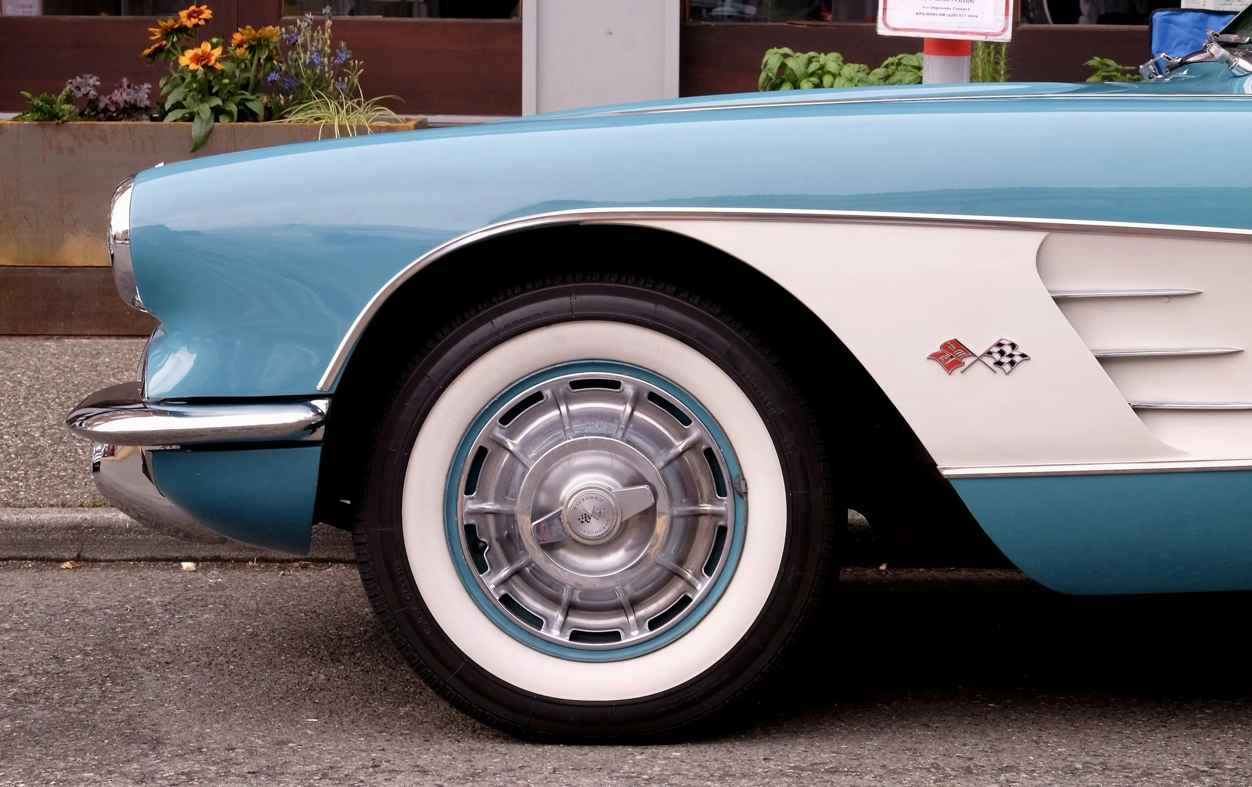 Close-up of the front left side of a vintage blue and white Chevrolet Corvette with whitewall tires, chrome hubcap, and Corvette insignia on the fender.