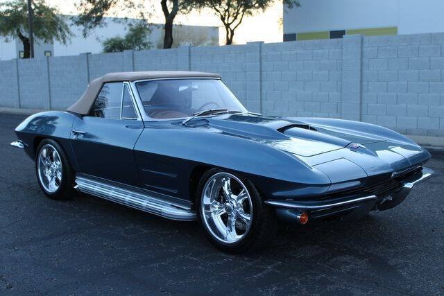 A classic dark blue Chevrolet Corvette convertible with chrome wheels parked on a paved lot near a concrete wall and trees.