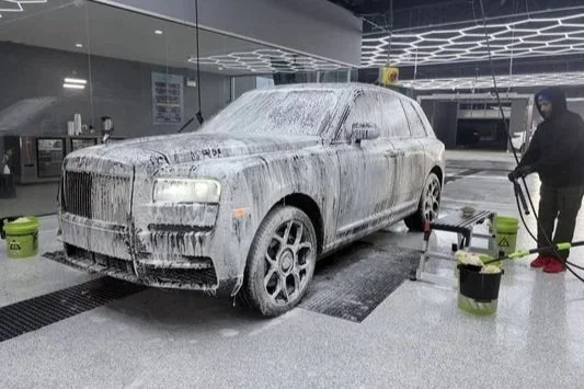 A luxury SUV at a car wash station covered in soap suds, with a worker cleaning the vehicle.