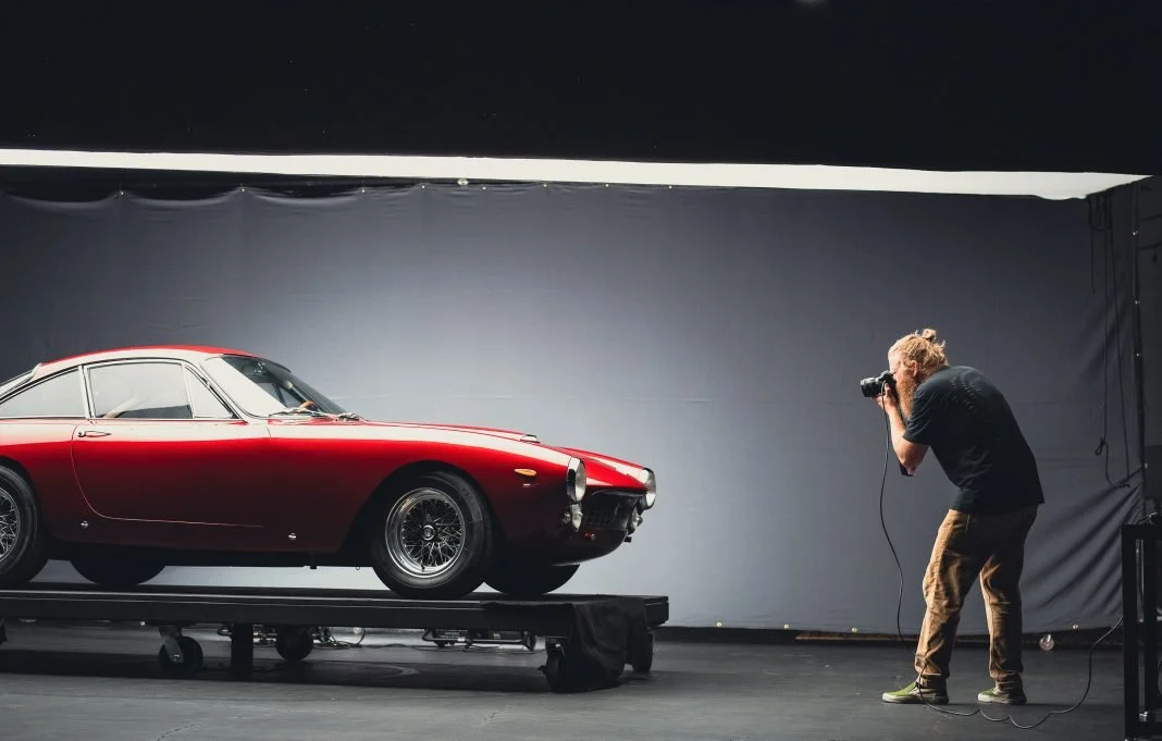 A photographer taking a picture of a vintage red sports car on a platform in a studio.