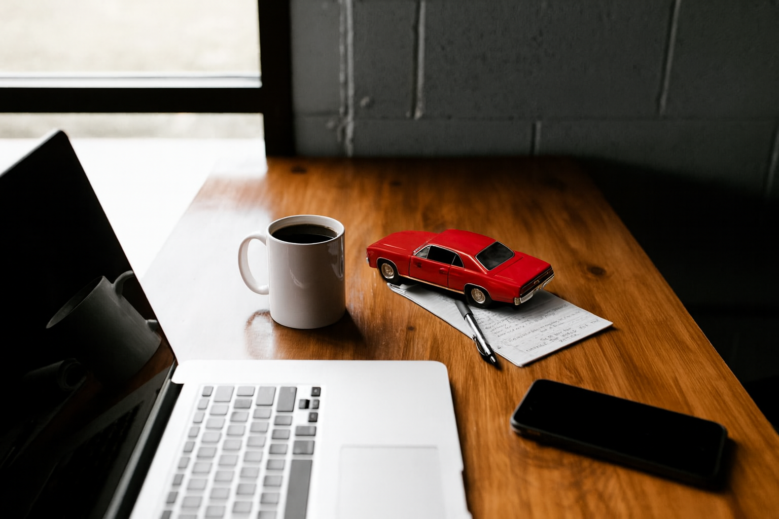 A wooden desk with a laptop, a smartphone, a white mug of coffee, a red toy car placed on a notepad with handwritten notes, and a pen.