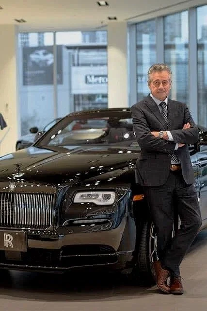 A man in a suit standing next to a black luxury car inside a modern building with large windows.
