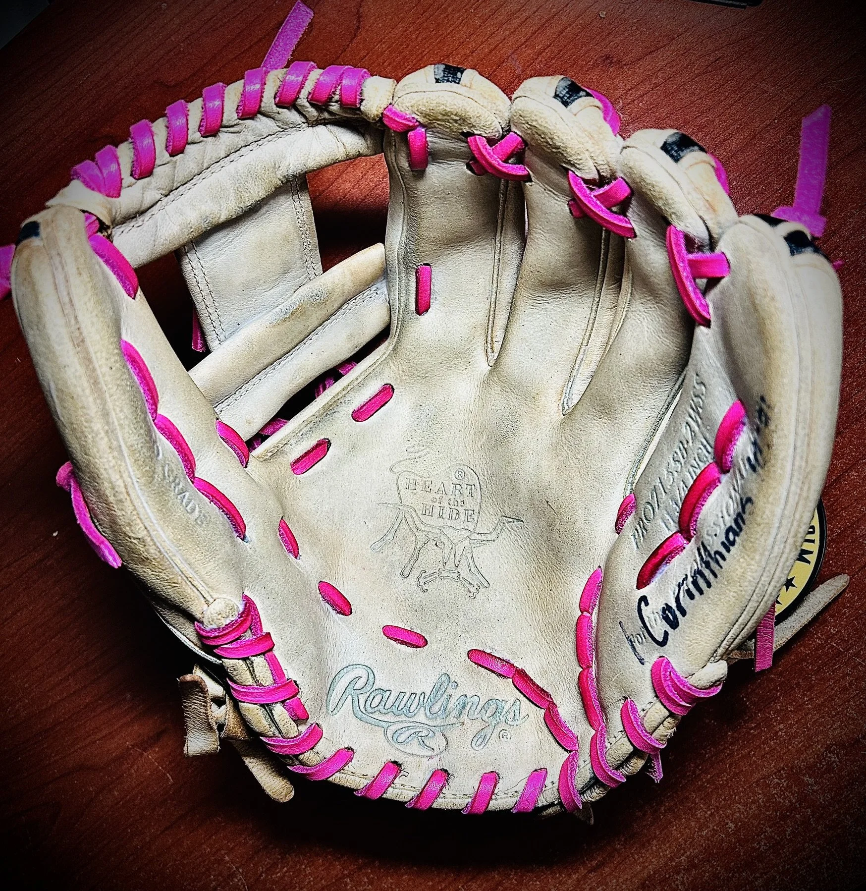 A worn white Rawlings baseball glove with pink lacing on a wooden surface.